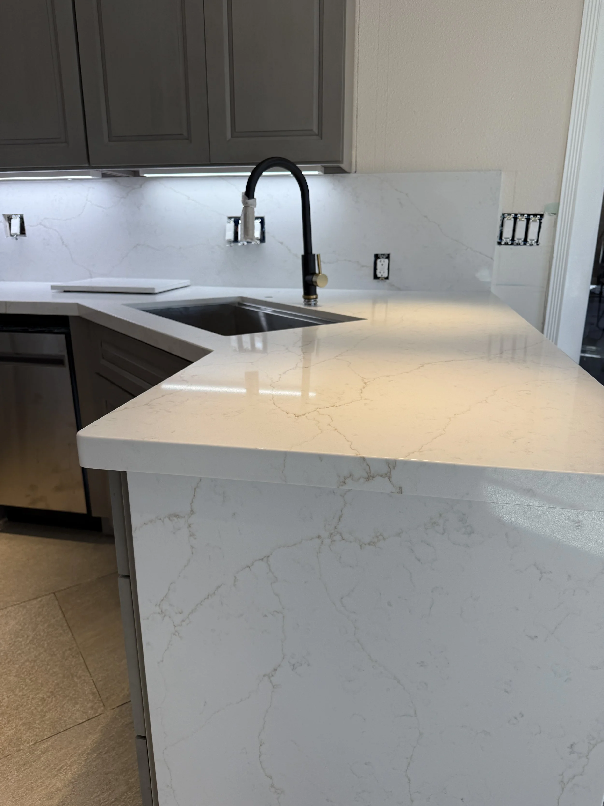 Kitchen with white marble countertops, dark gray cabinets, and a black faucet over a stainless steel sink, with some wall outlets and switches under installation.