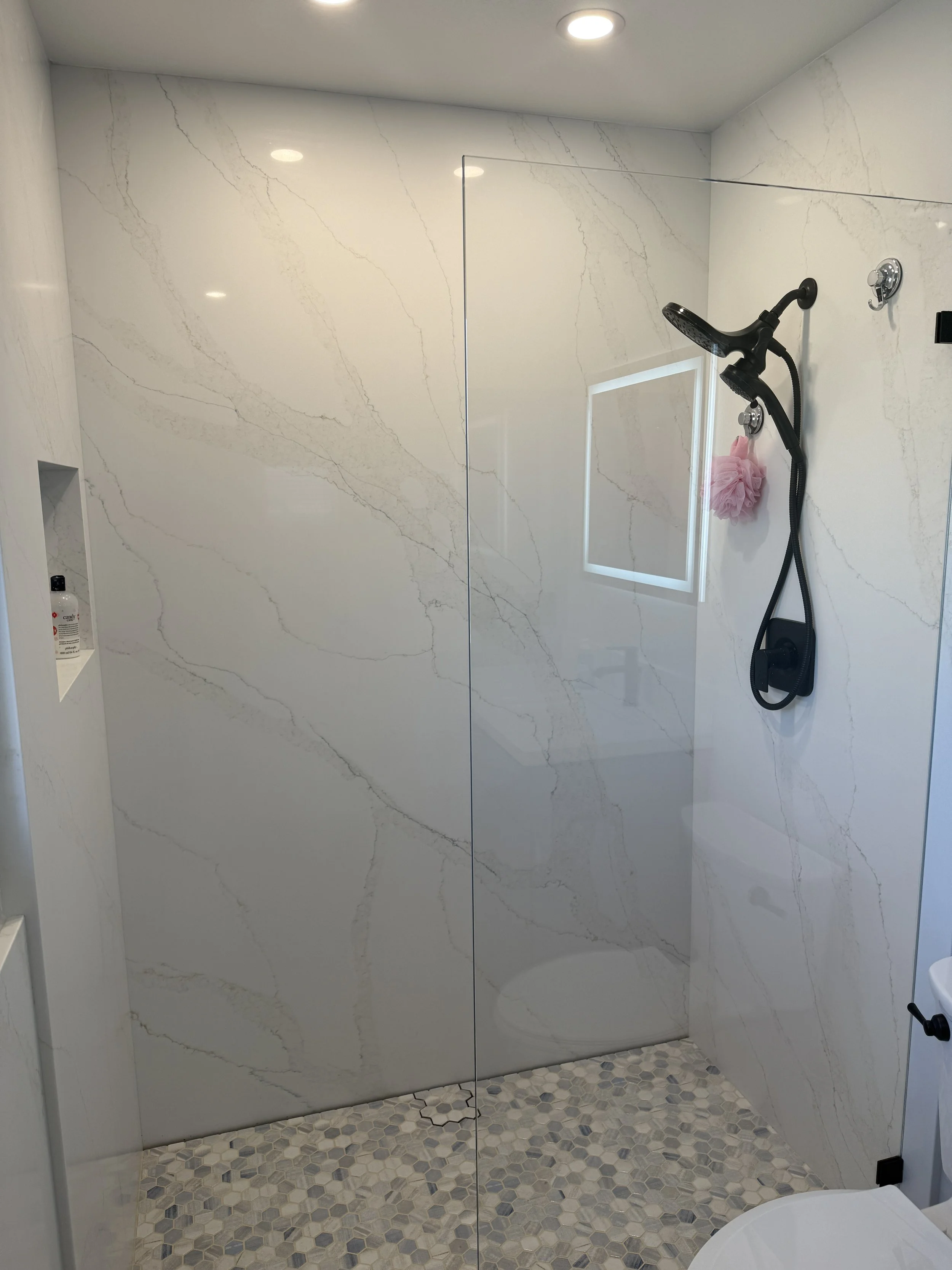 Modern walk-in shower with marble-look walls, pebble tile floor, black handheld showerhead, pink shower puff, and a small built-in shelf with toiletries.