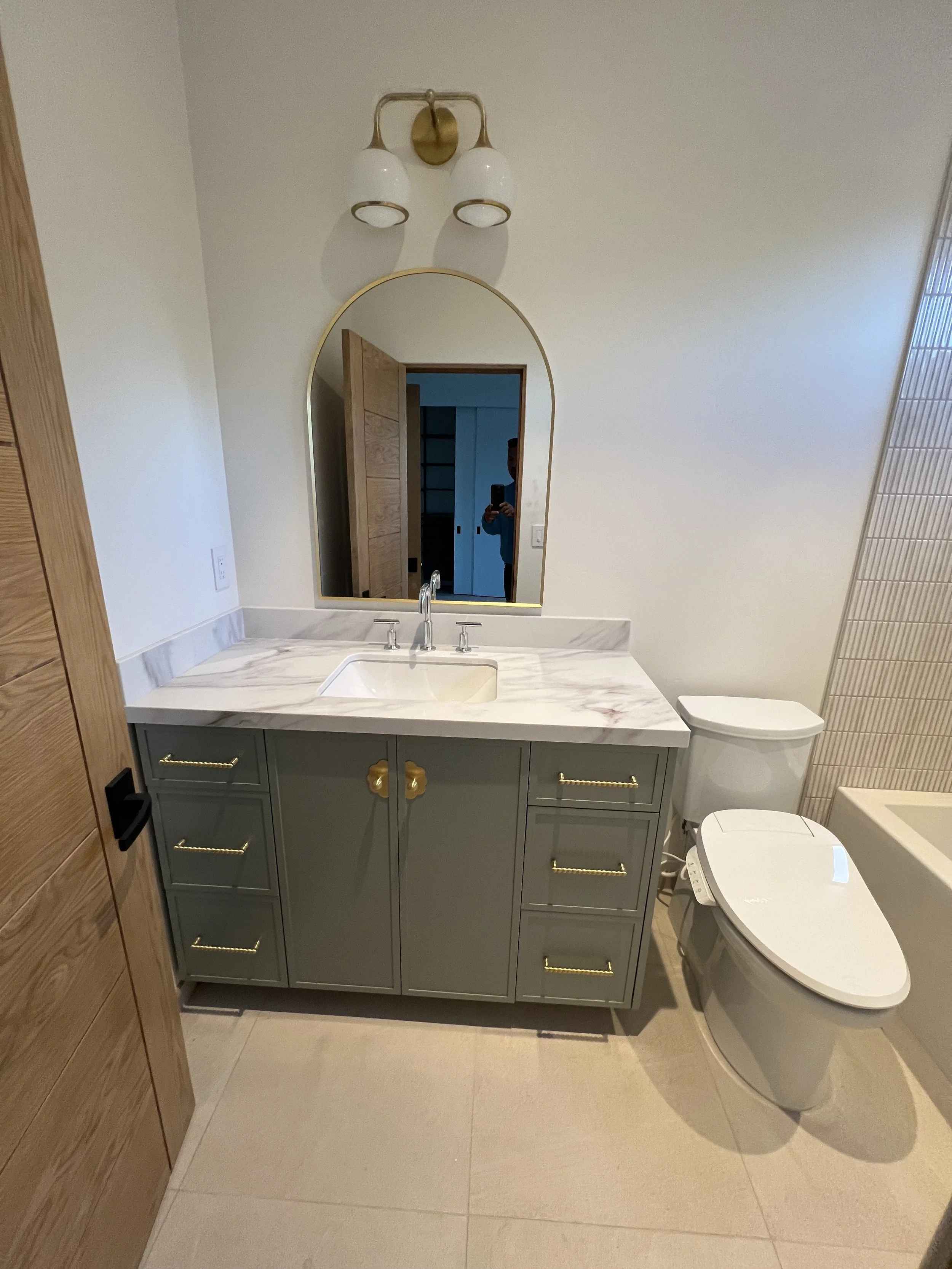 Bathroom with white walls, a vanity with a marble countertop, green cabinets with gold handles, a large mirror with rounded top, a wall-mounted light fixture, a toilet, and a tiled shower.