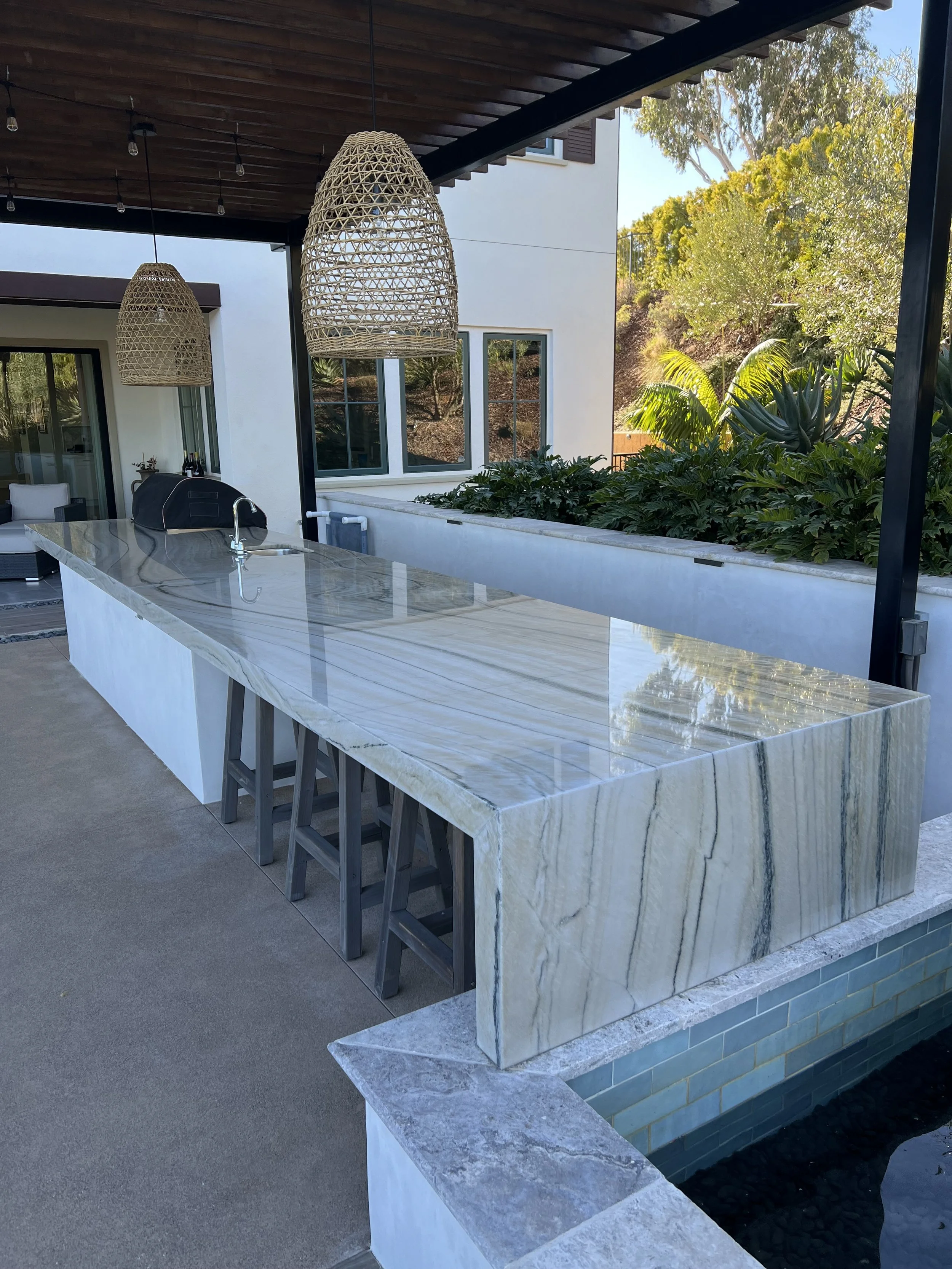 Outdoor kitchen with a large marble countertop, three stools underneath, hanging wicker pendant lights, and greenery in the background.