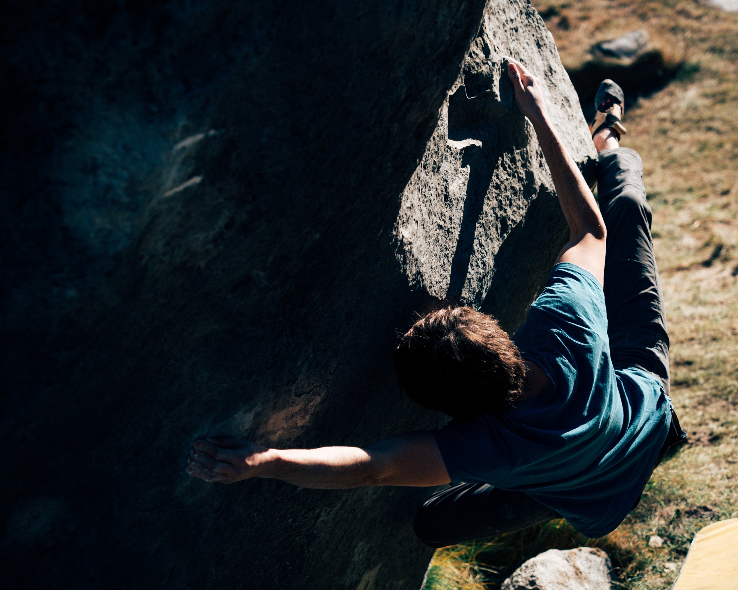A man engages in bouldering, reaching for a hold on an outdoor rock face, wearing a blue shirt and black shorts.