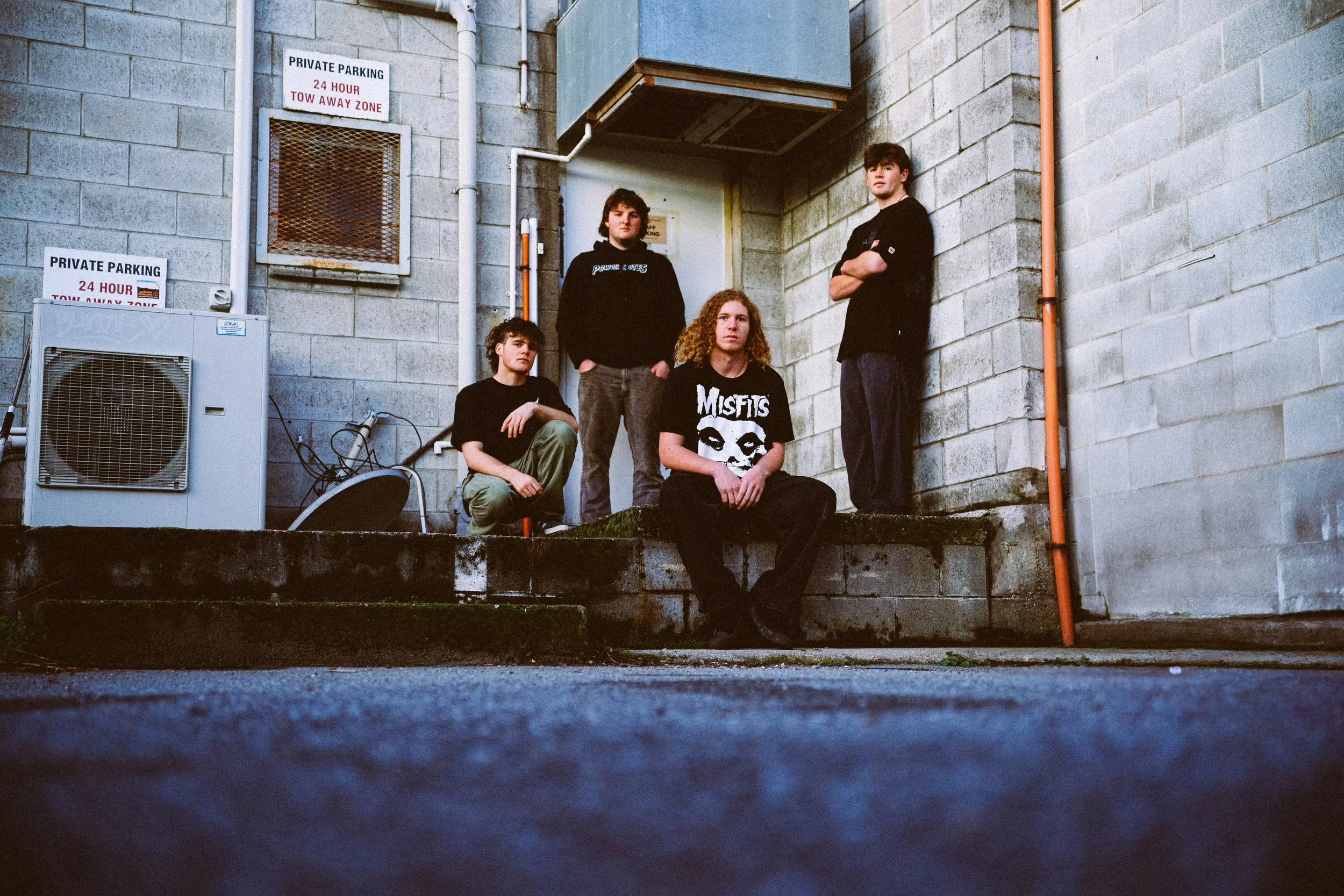 Four young people with curly and straight hair posing against a concrete wall outside, near signs that read 'Private Parking 24 Hour Tow Away Zone.' One person is sitting on the ledge, while others stand or sit nearby, all dressed casually.