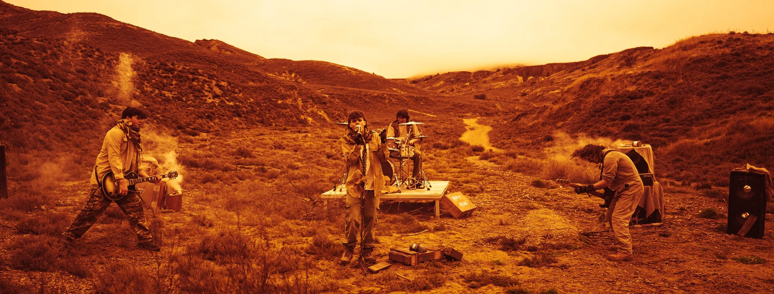 A band performing outdoors on a dirt trail in a hilly landscape with orange-tinted sky, including a guitarist, a drummer, and a vocalist with microphones.