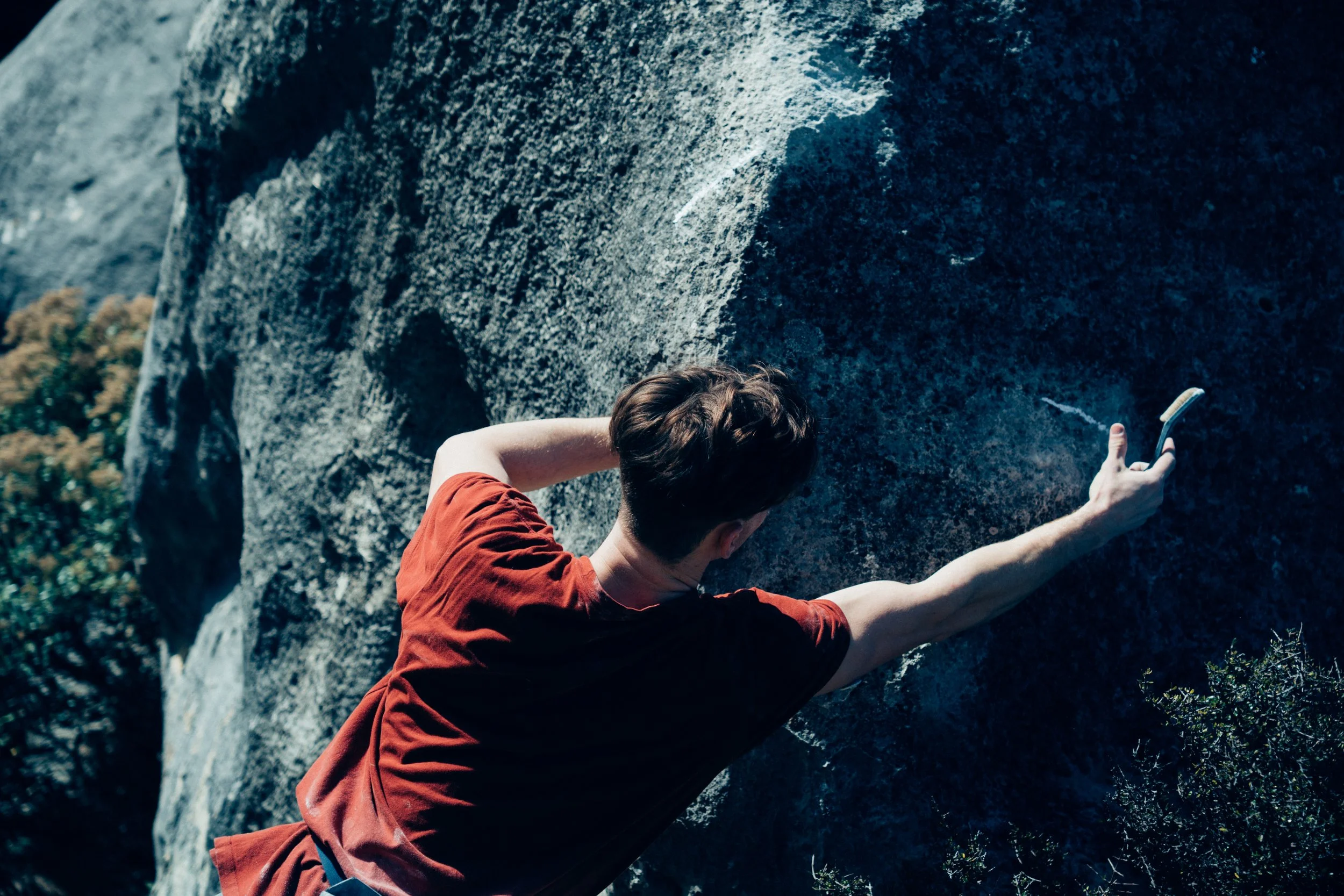 A person in a red shirt rock climbing on a large rock face with a harness visible at the waist, holding a smartphone in their extended arm.