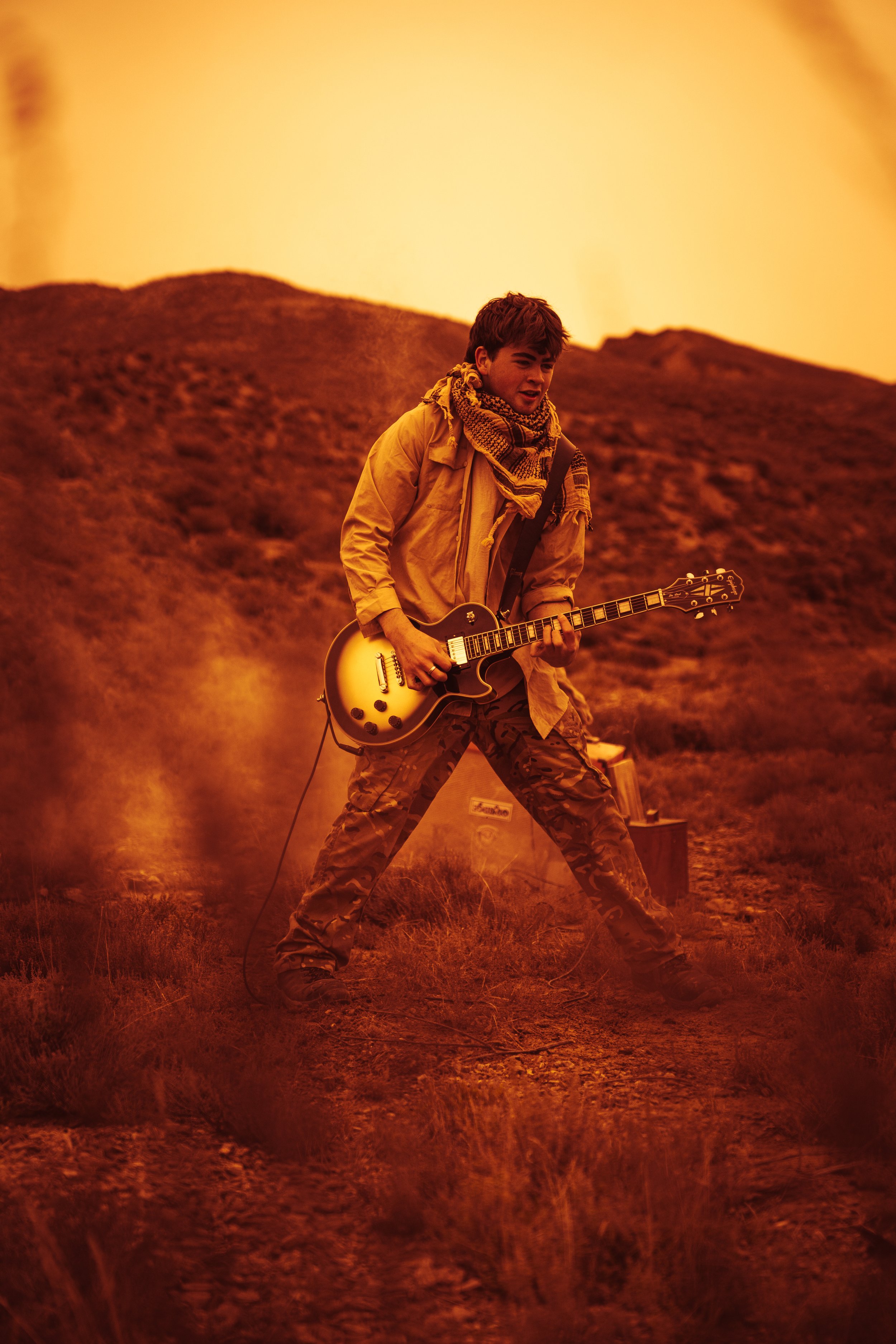 A young man playing an electric guitar outdoors during sunset, wearing a yellow jacket, camouflage pants, and a scarf, with a hill in the background and equipment nearby.