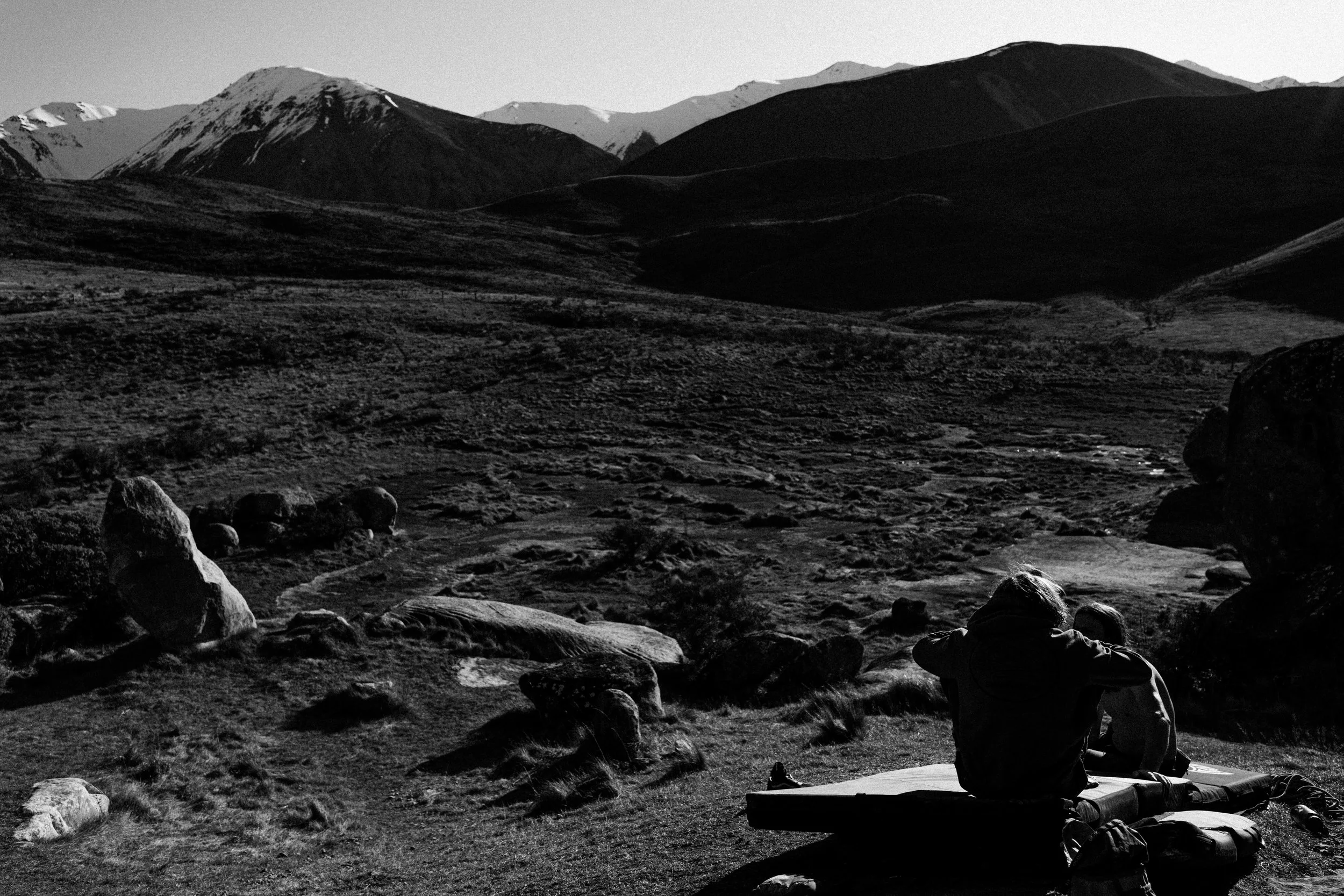 A black and white photograph of a mountainous landscape with two people sitting on a flat surface, possibly a mat or blanket, observing the view.