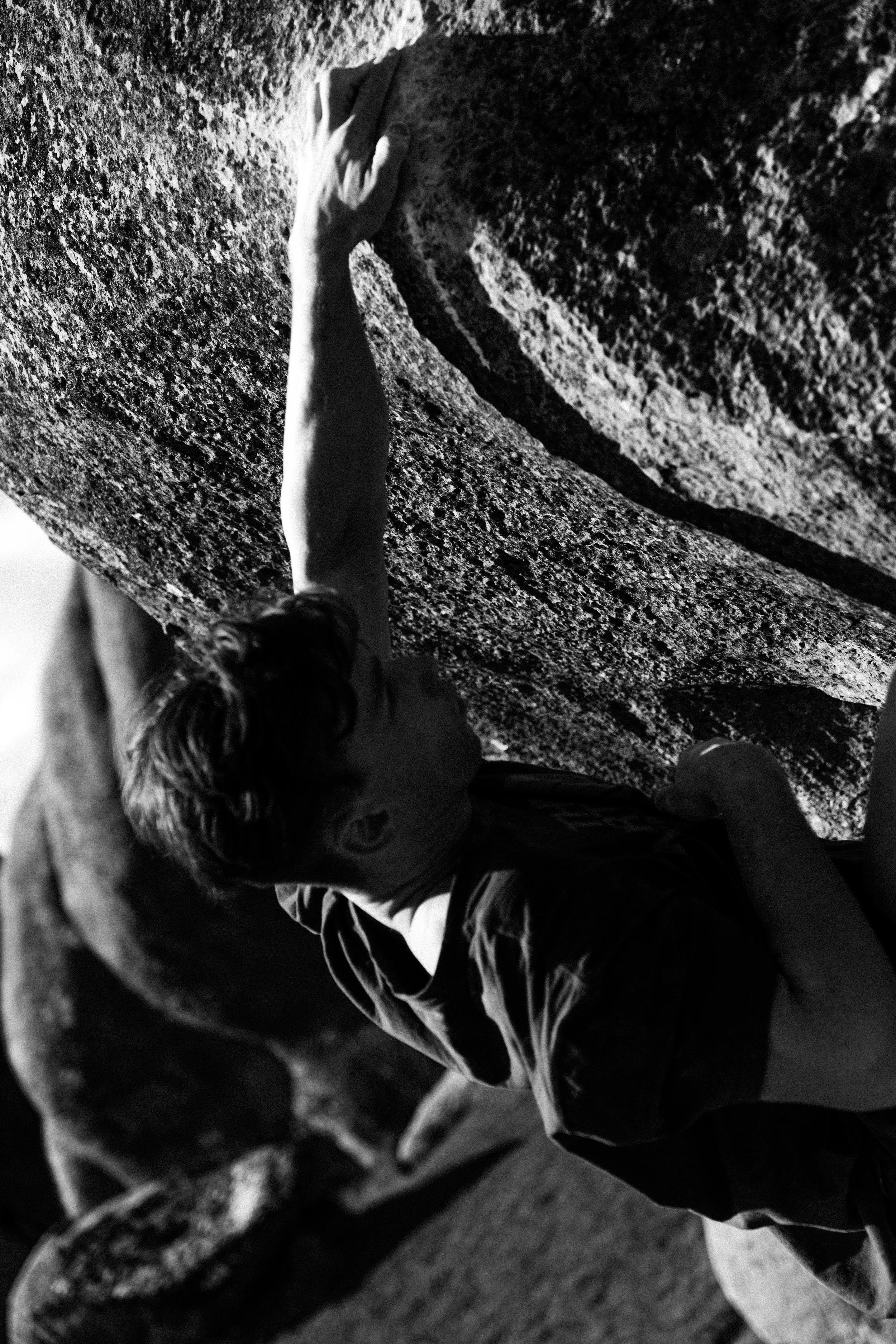 A person climbing a large, textured boulder outdoors, reaching upward with one hand, in black and white.