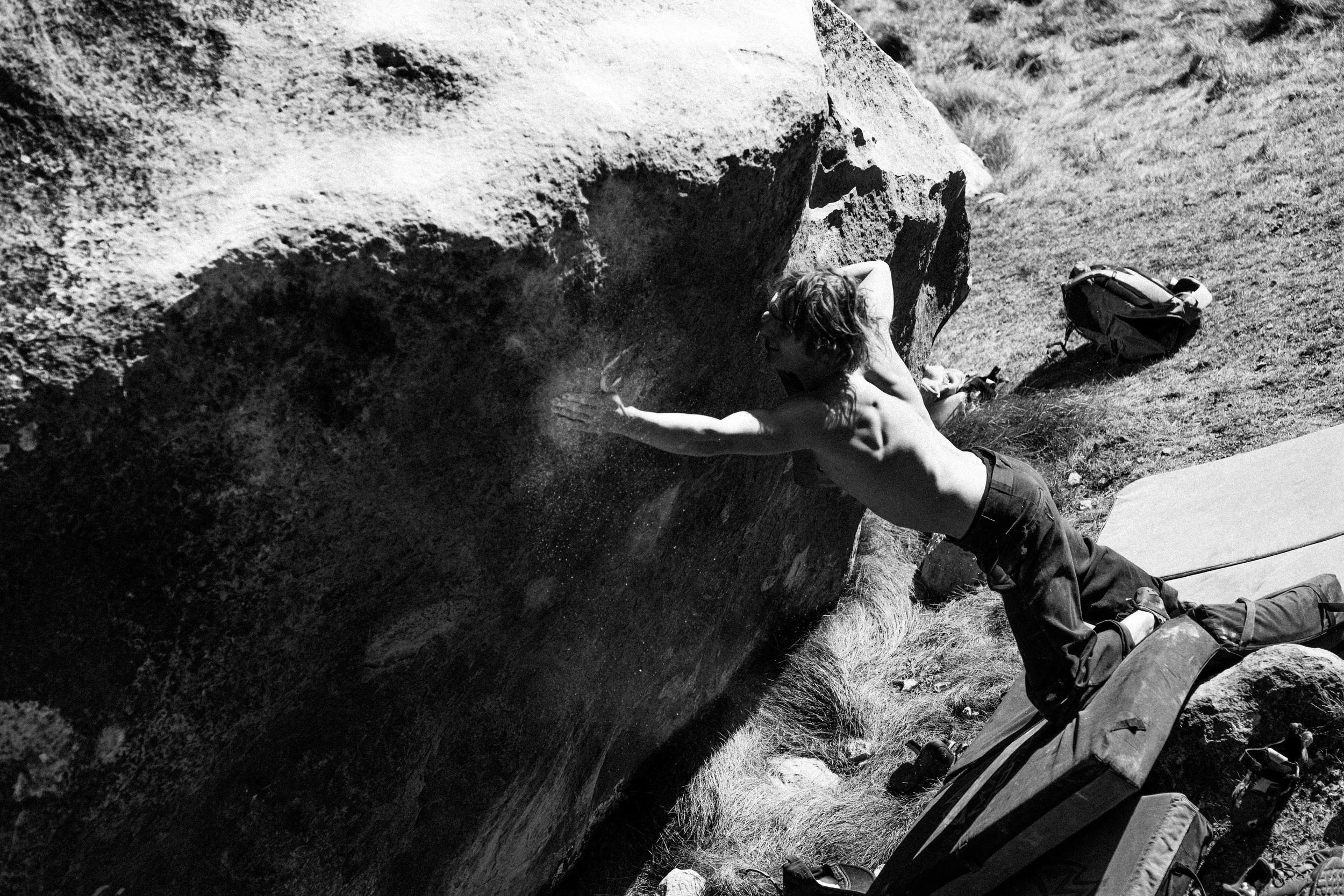 A person with long hair and no shirt bouldering on a large rock outdoors, with climbing gear and a bag nearby.
