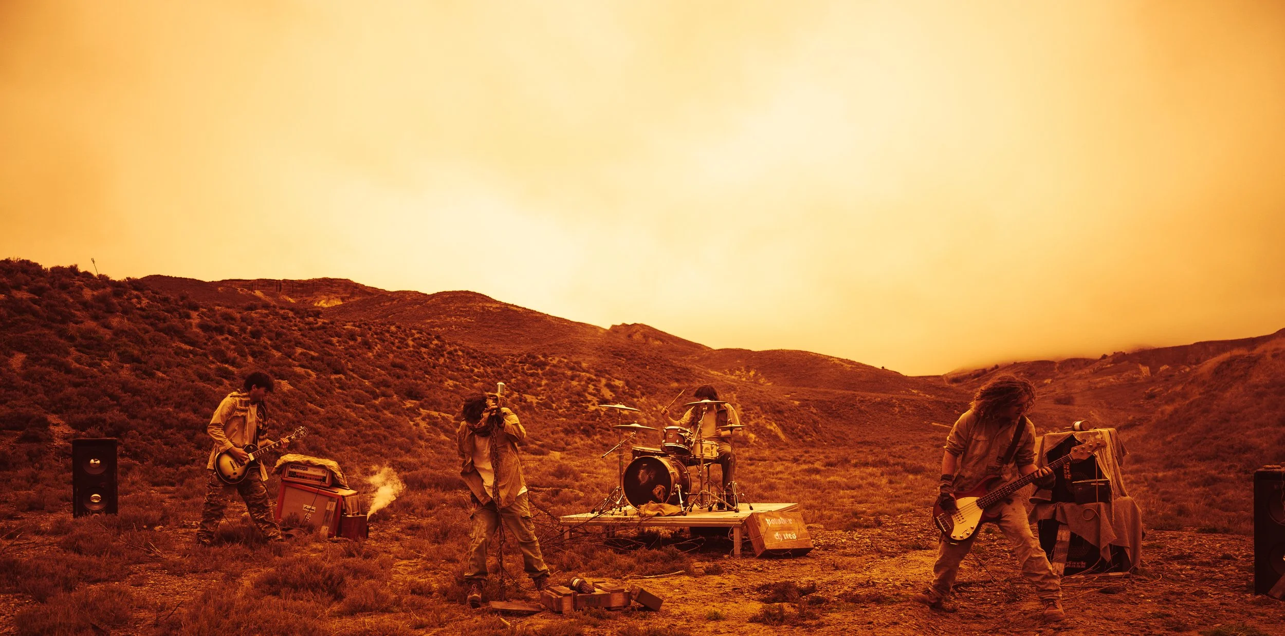 A band performing outdoors on a stage in a desert landscape at sunset with mountains in the background, playing guitars and drums.