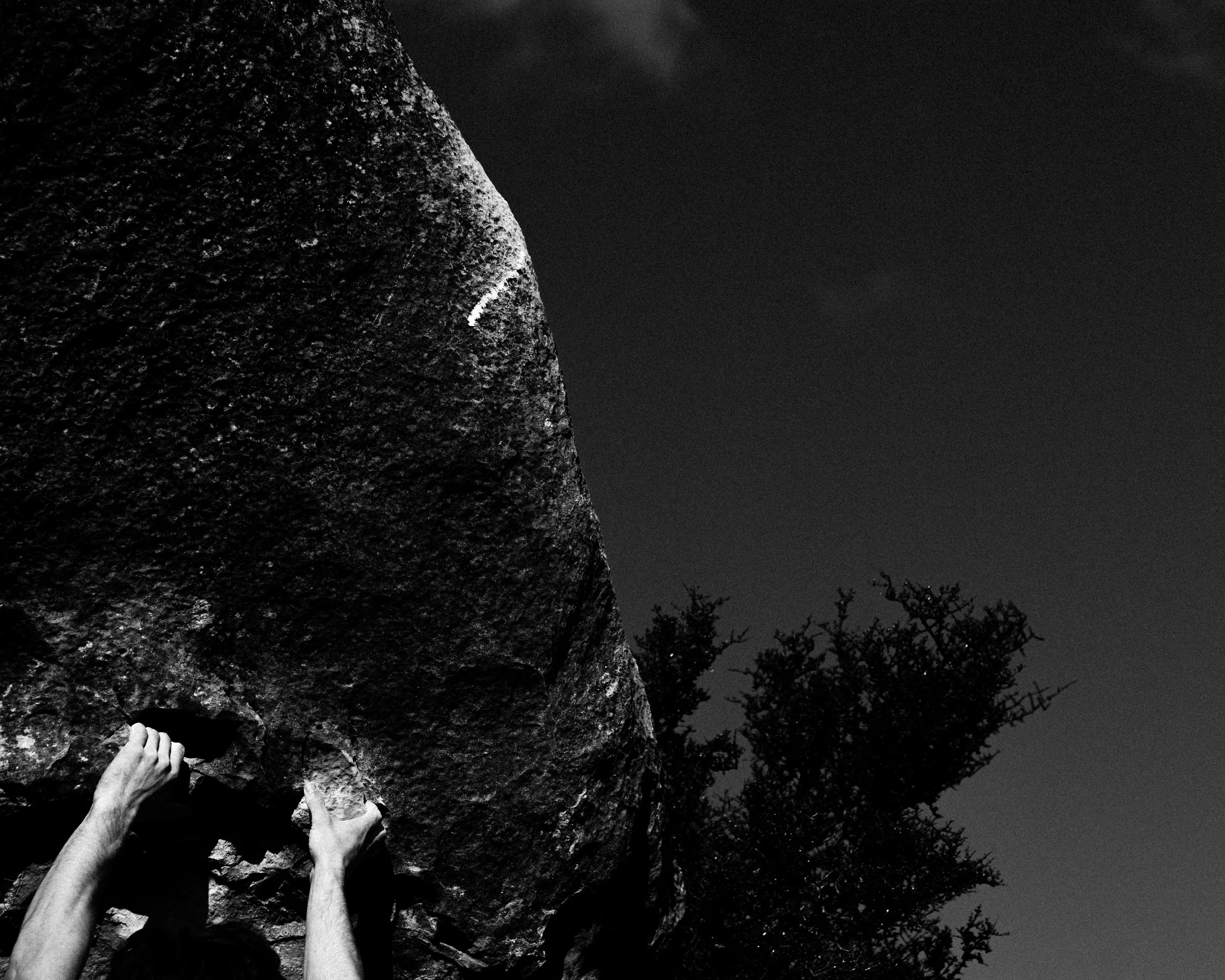 Hands reaching to climb a large rock formation outdoors at night with trees in background.