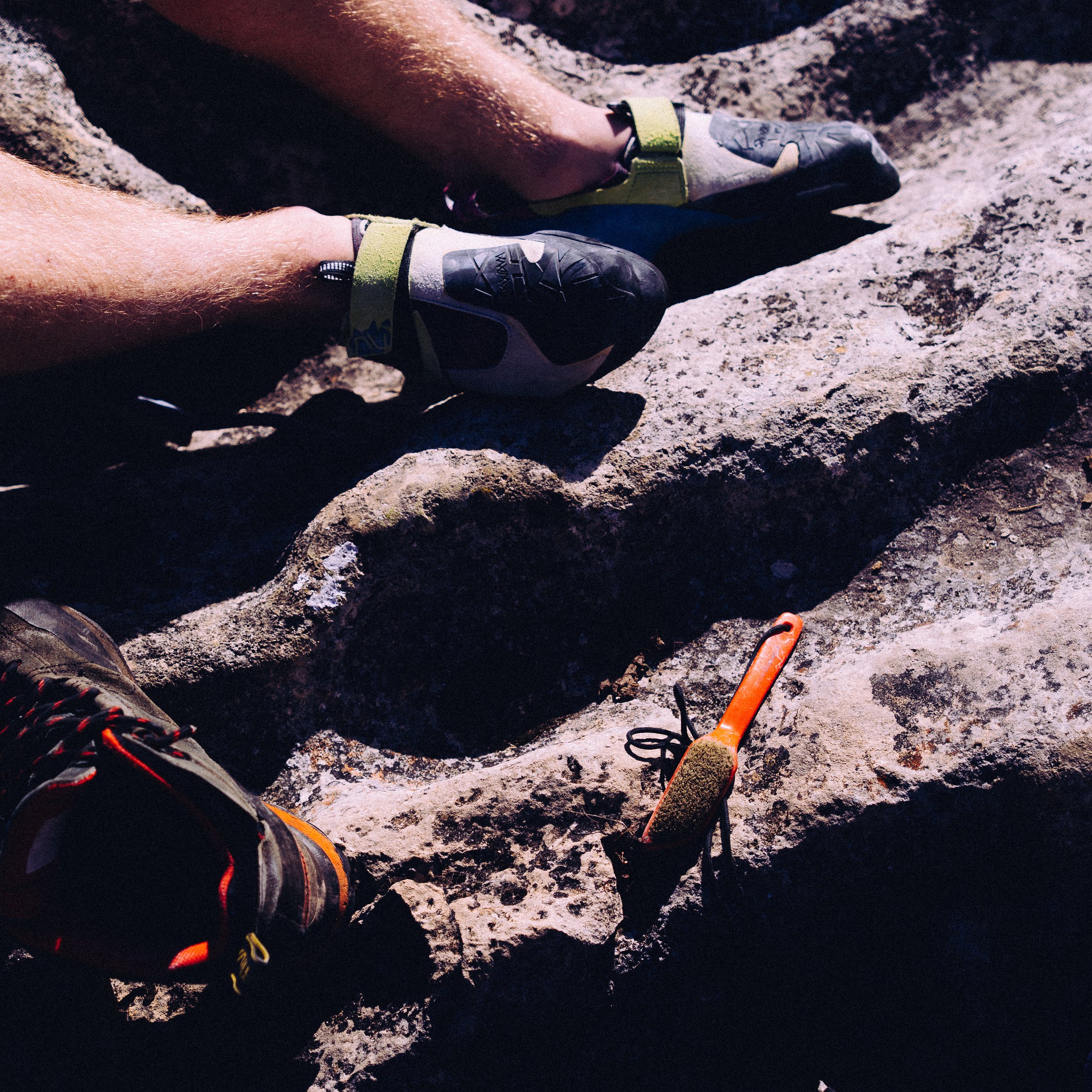 Close-up of rock climbers' feet with climbing shoes and a gear tool on the rock surface.
