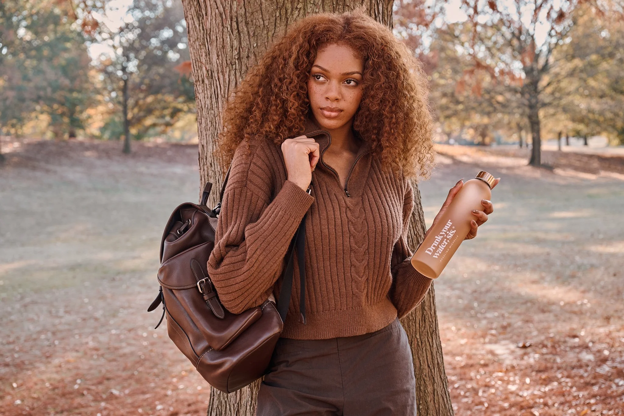A young woman with curly hair leaning against a tree in a park during autumn, holding a water bottle and carrying a brown backpack.