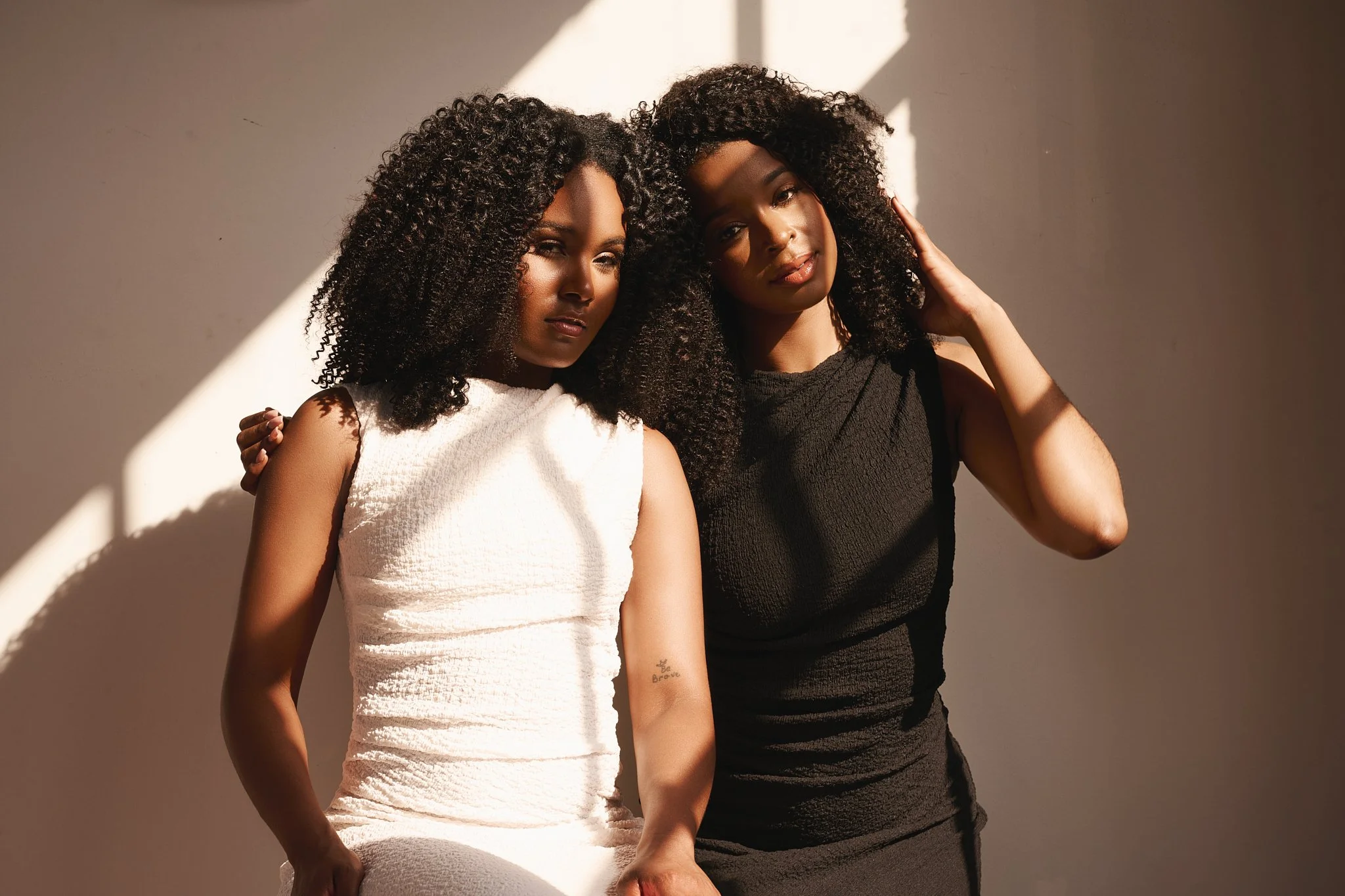 Two women with curly hair posing against a neutral background with shadows and lighting.