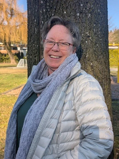 A smiling woman wearing glasses, a light gray puffer jacket, a green top, and a gray scarf, standing outdoors next to a tree with a park background.