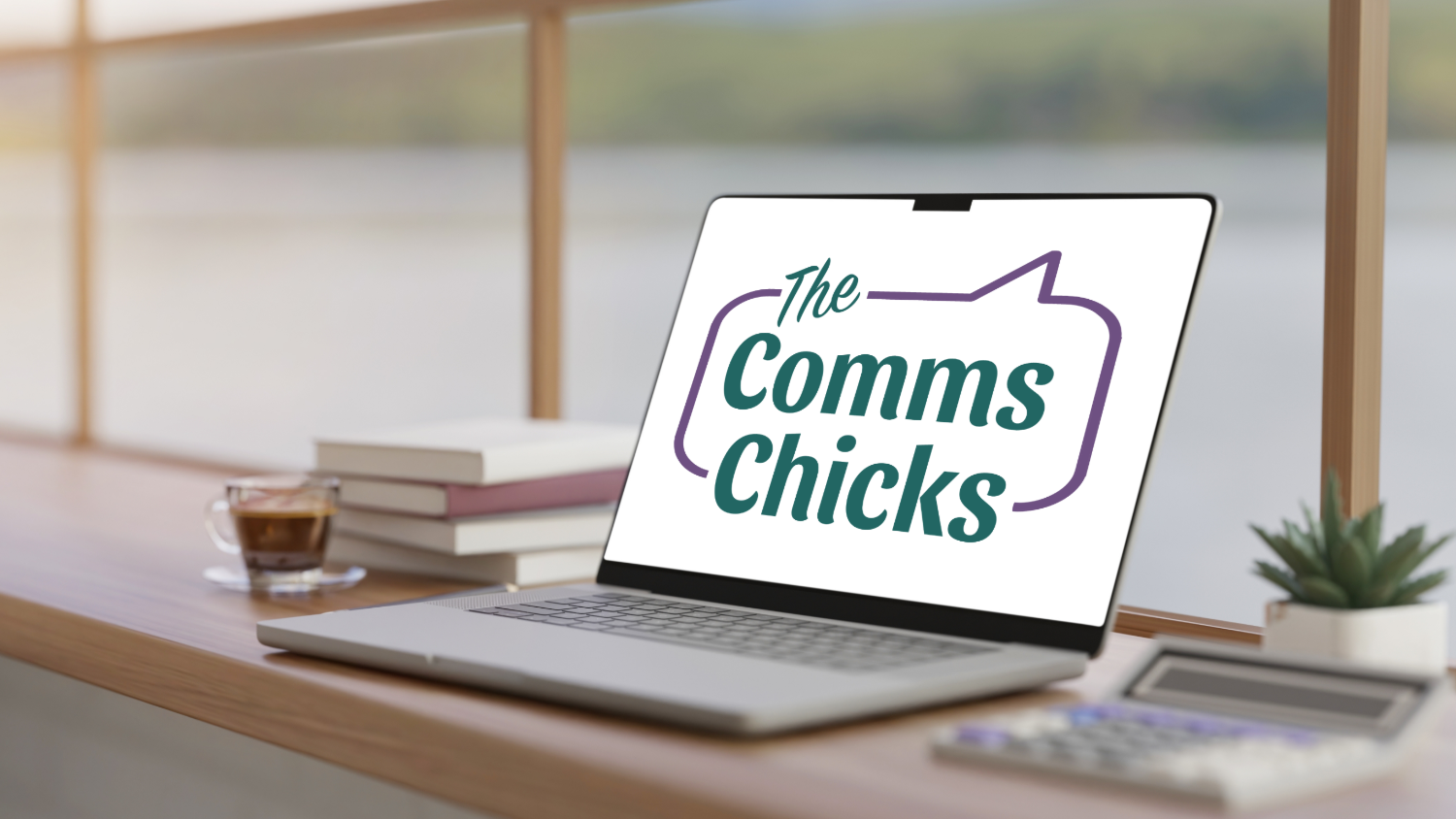 A laptop on a wooden desk displays the logo 'The Comms Chicks'. The desk has a stack of books, a cup of coffee, a potted plant, a calculator, and a blurred background of a window with a scenic view.