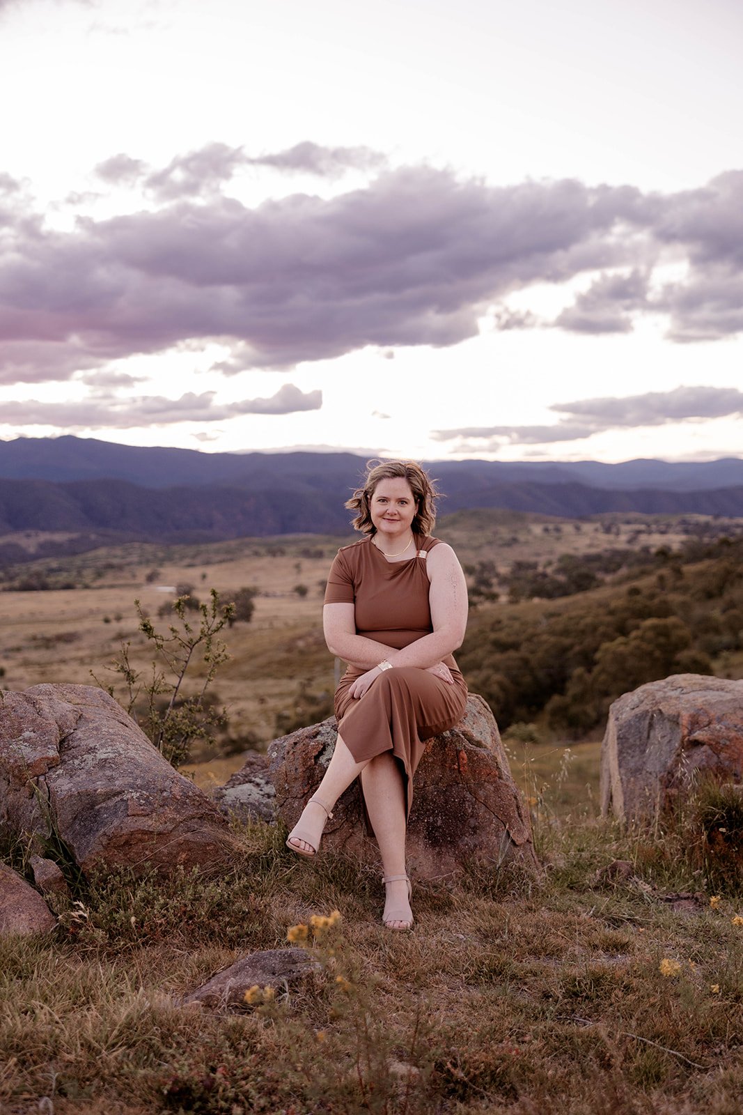 A woman sitting on a rock in a desert landscape with mountains and cloudy sky in the background.