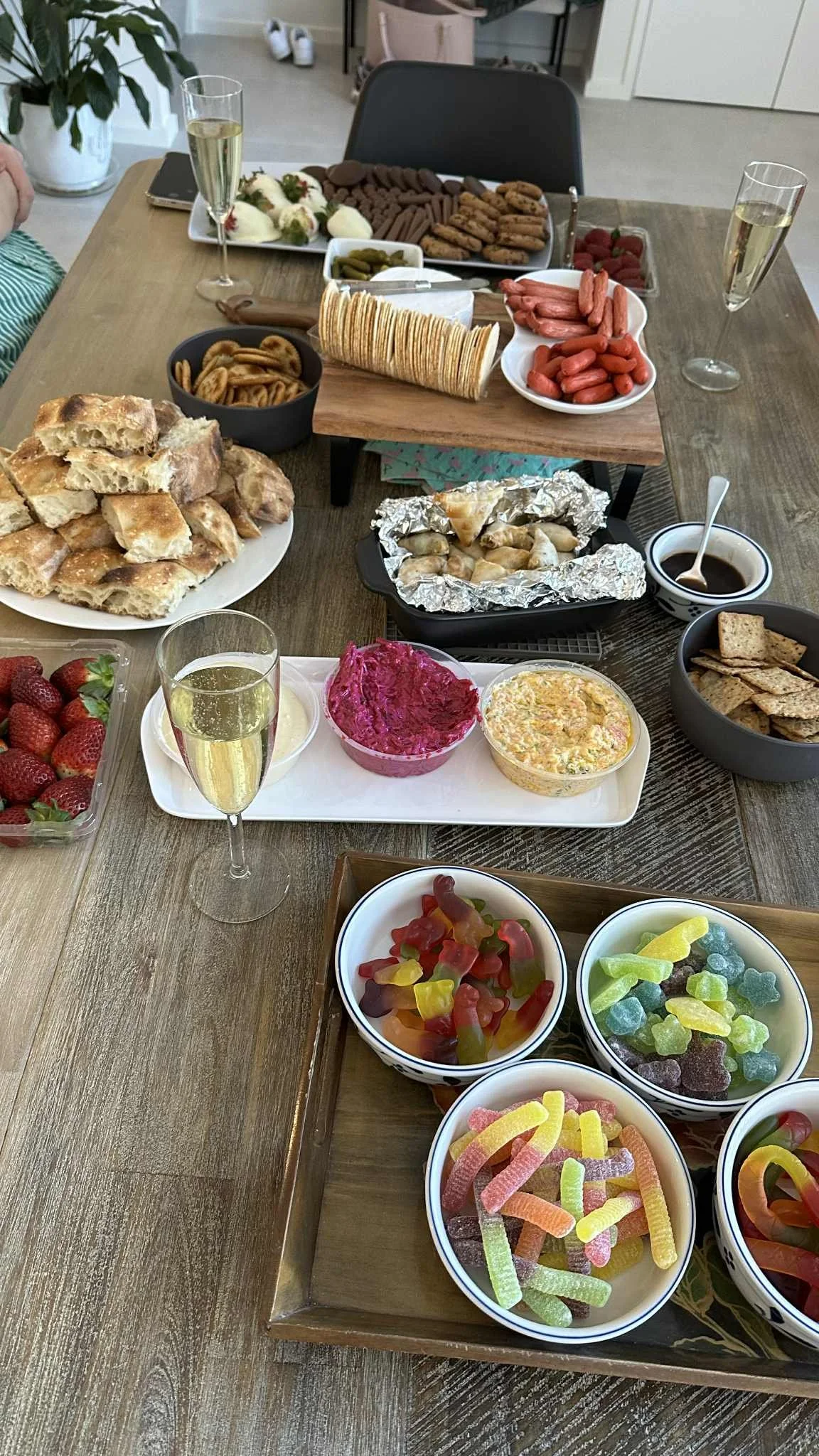 A table set with party snacks including bread, dips, crackers, sausages, lollies, strawberries, biscuits, and glasses of champagne.