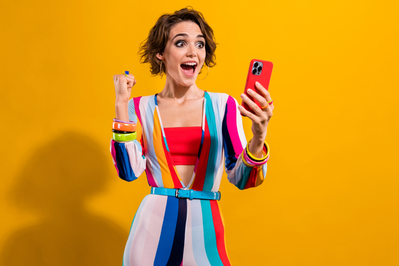 Woman in colorful striped outfit celebrating while looking at her phone against a yellow background.