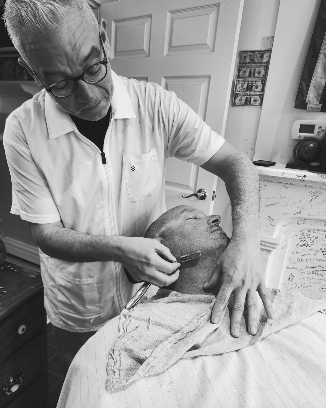 Black and white photo of Steve R shaving for a client who sits in the chair