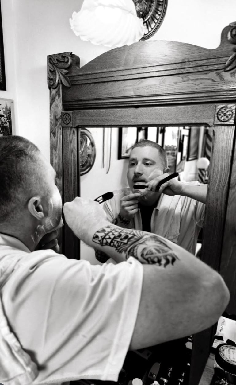 Black and white photo of Conner Woodring shaving for himself in the mirror at Horse and Lion Barber Co.