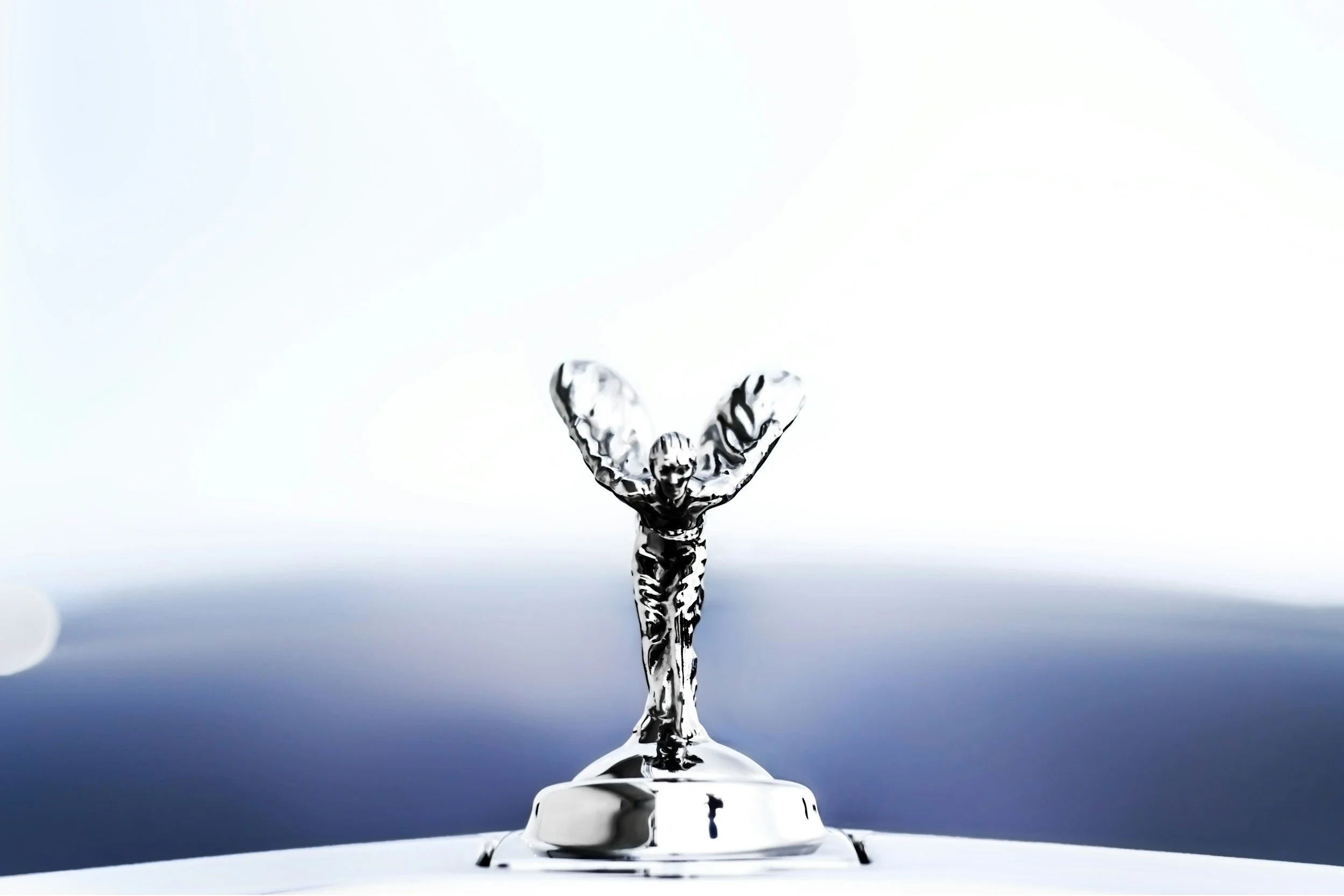 Close-up of a silver car hood ornament depicting a figure with wings, set against a blue and white sky.