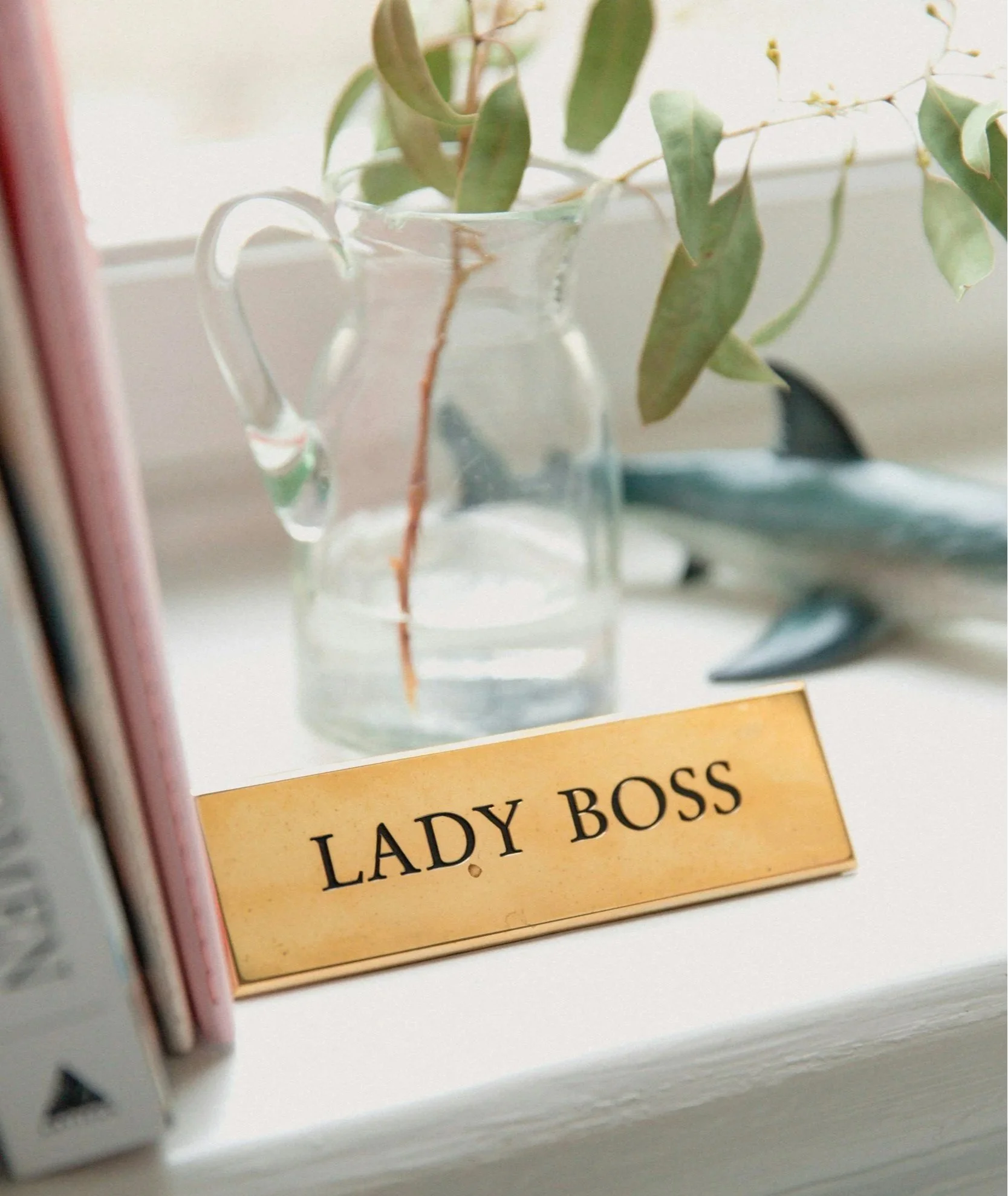 Close-up of a nameplate reading 'Lady Boss' placed on a white surface, with a glass pitcher containing green leaves in the background.