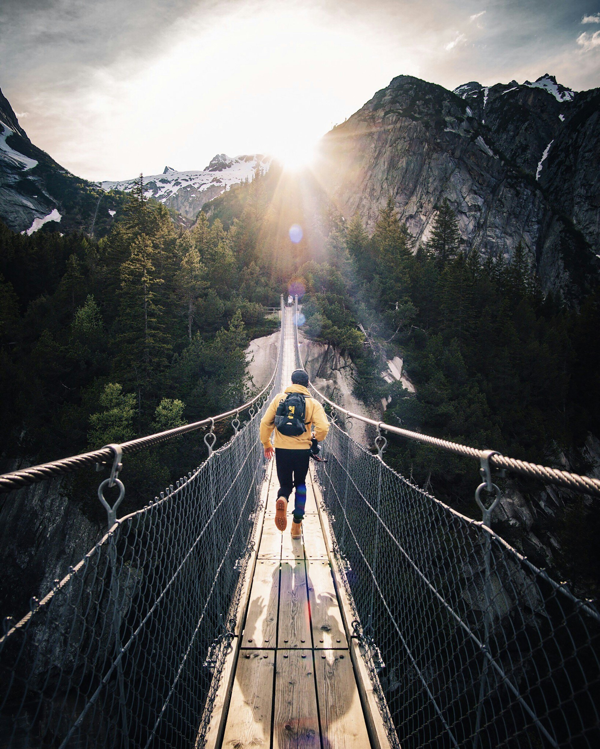 Person walking across a suspension bridge in a mountainous forest area during sunset.