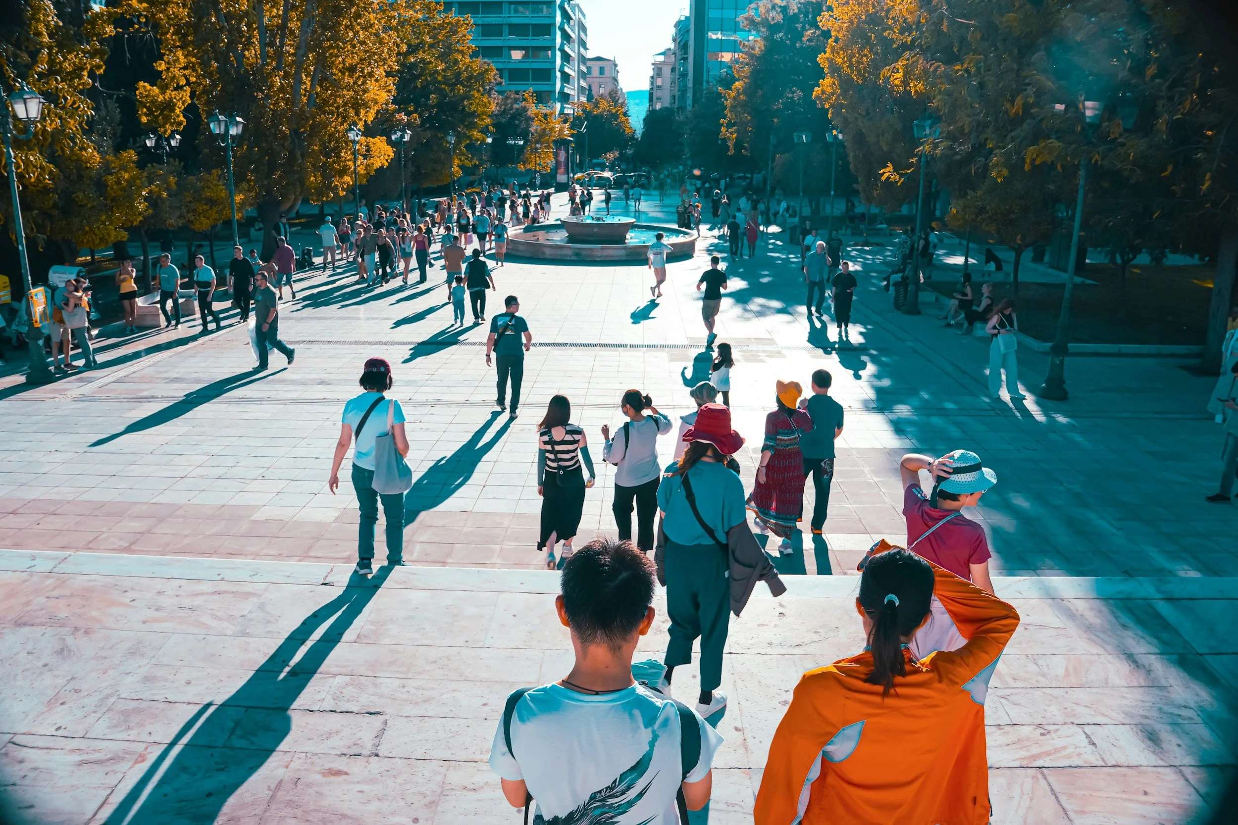 People walking in a city square with trees, benches, and a fountain, during late afternoon.