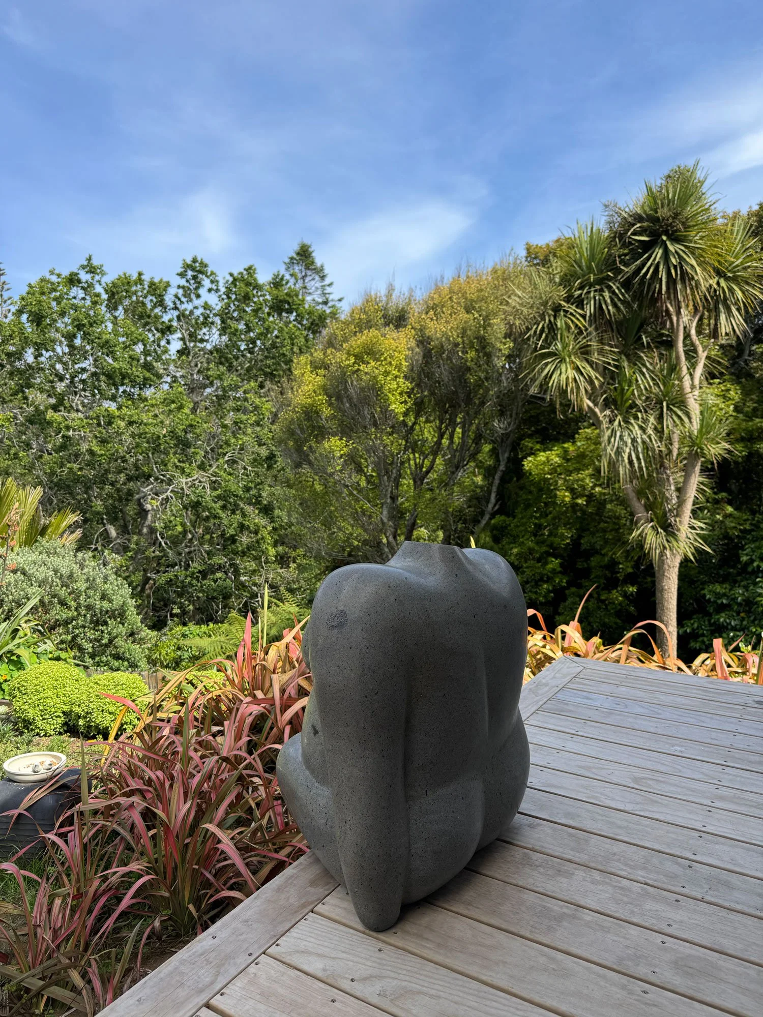 A black stone sculpture of a human torso on a wooden deck surrounded by pink and green plants, with a lush green forest and blue sky in the background.