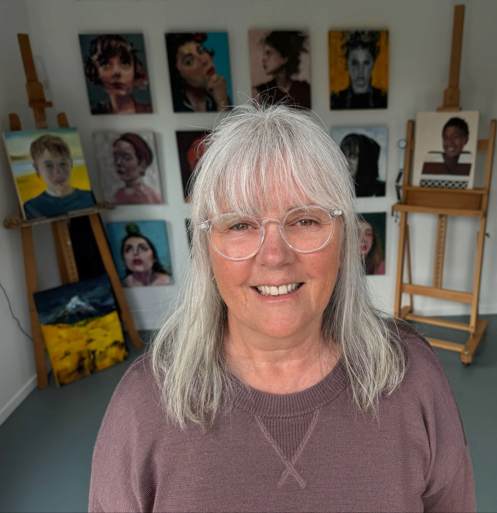 A woman with gray hair and glasses smiling in an art gallery with colorful portraits and paintings on easels behind her.