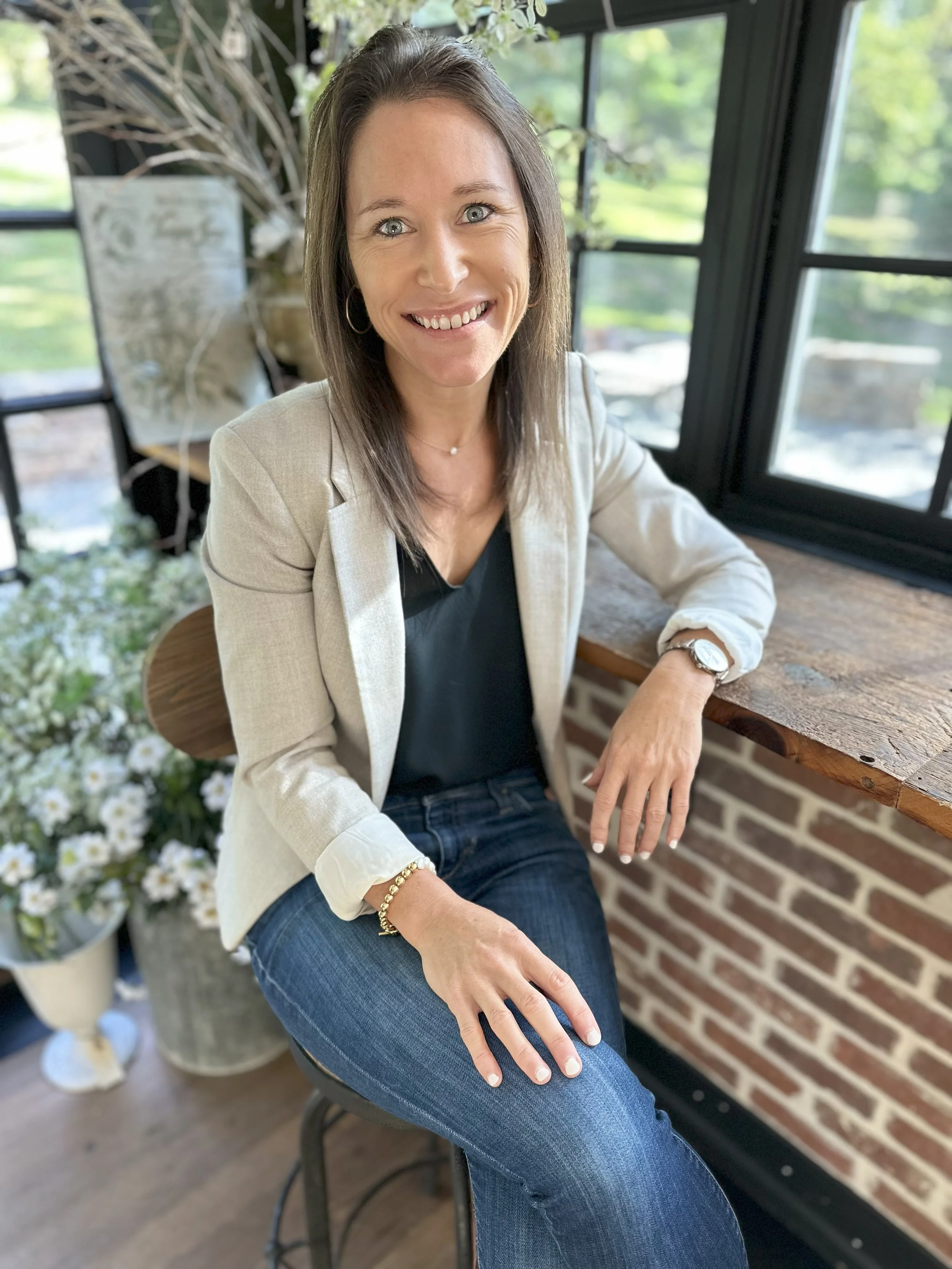 A woman with shoulder-length brown hair, blue eyes, and wearing a beige blazer, black top, and blue jeans, smiling and sitting on a stool in a bright room with large windows, a wooden windowsill, a brick wall, and potted plants in the background.