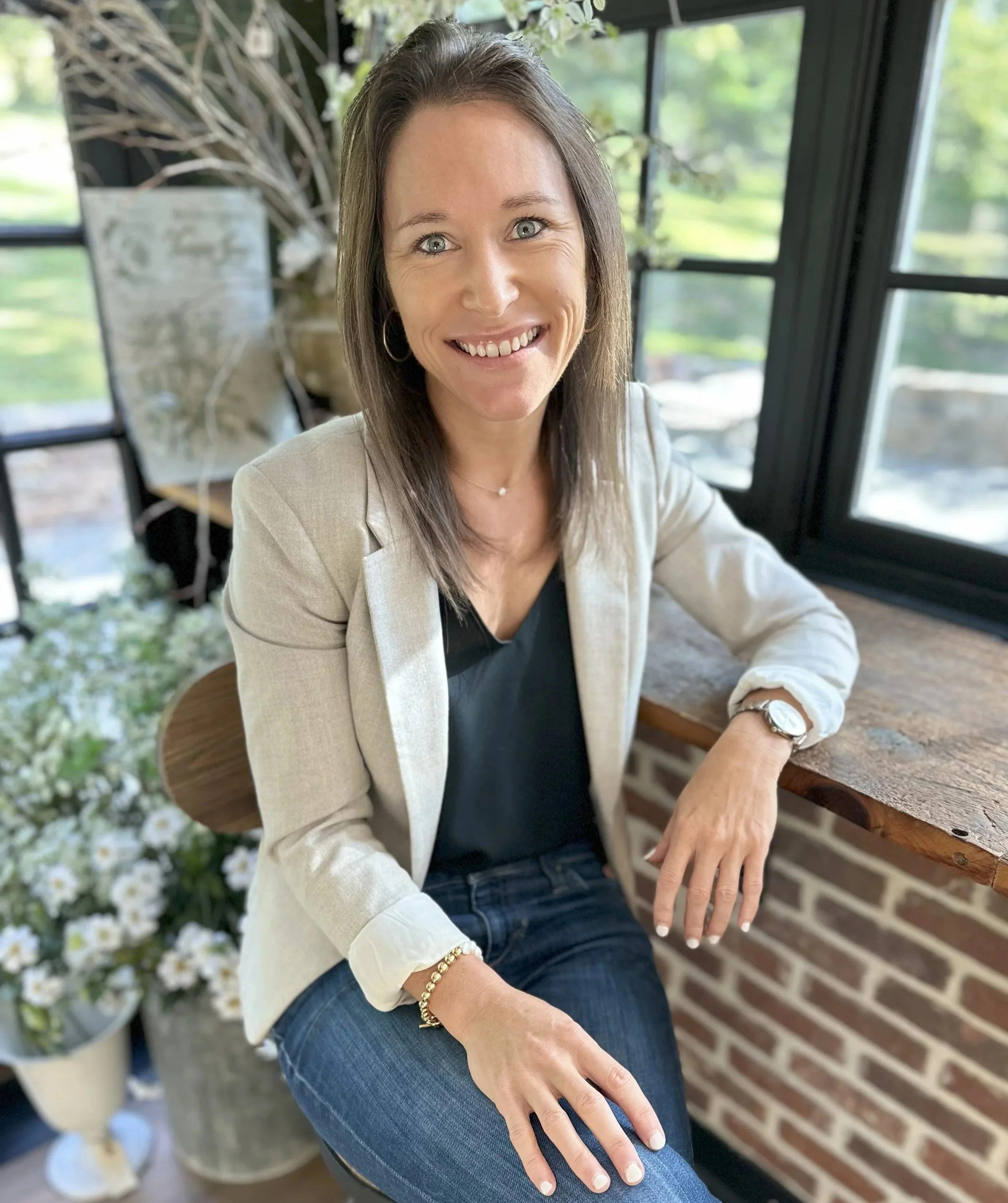 A woman with shoulder-length brown hair, blue eyes, wearing a beige blazer, black top, jeans, a watch, a bracelet, and hoop earrings, sitting at a wooden desk near large windows with a view of greenery outside, smiling at the camera.