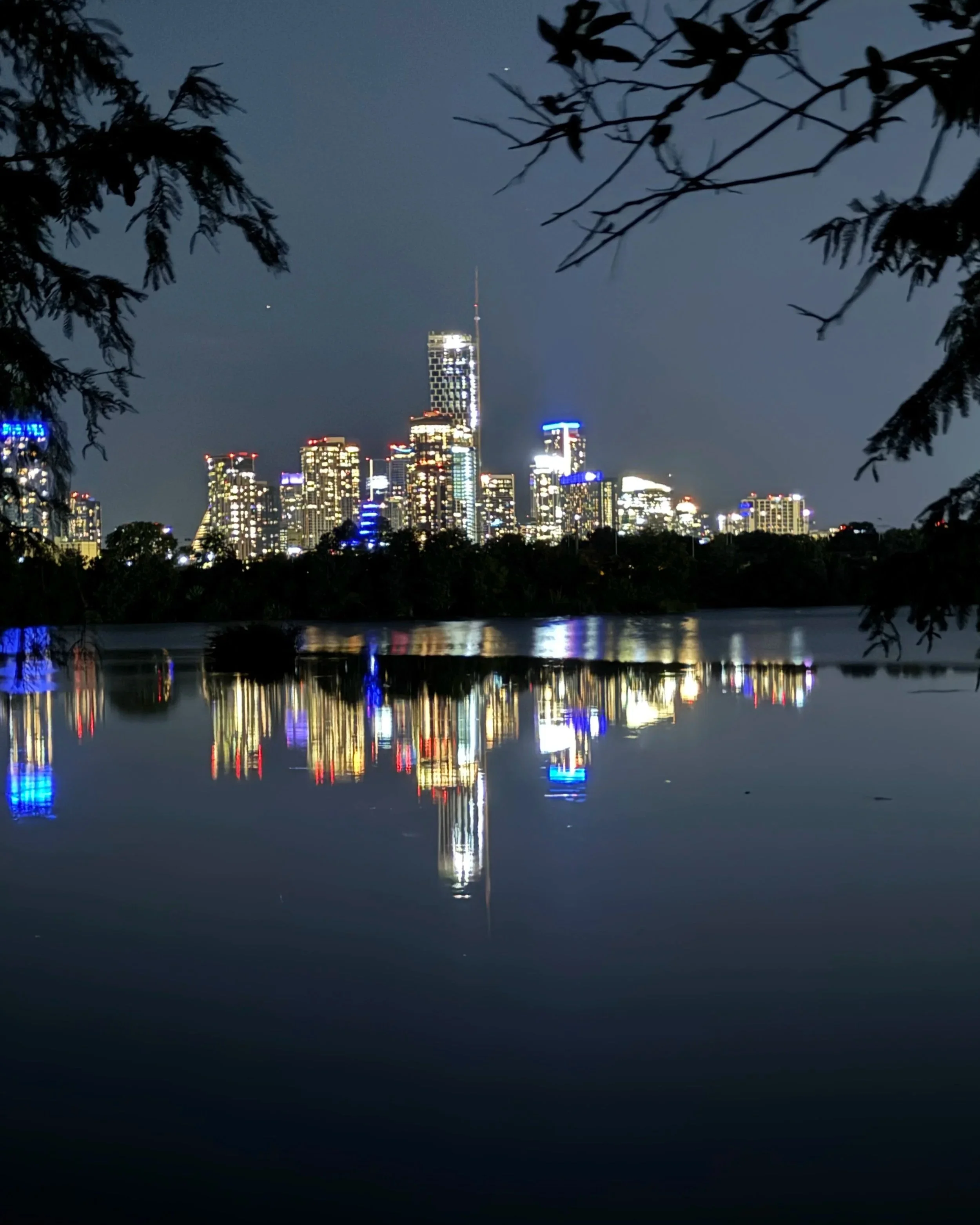 Nighttime Austin skyline with illuminated buildings reflecting on a Ladybird Lake, framed by tree branches