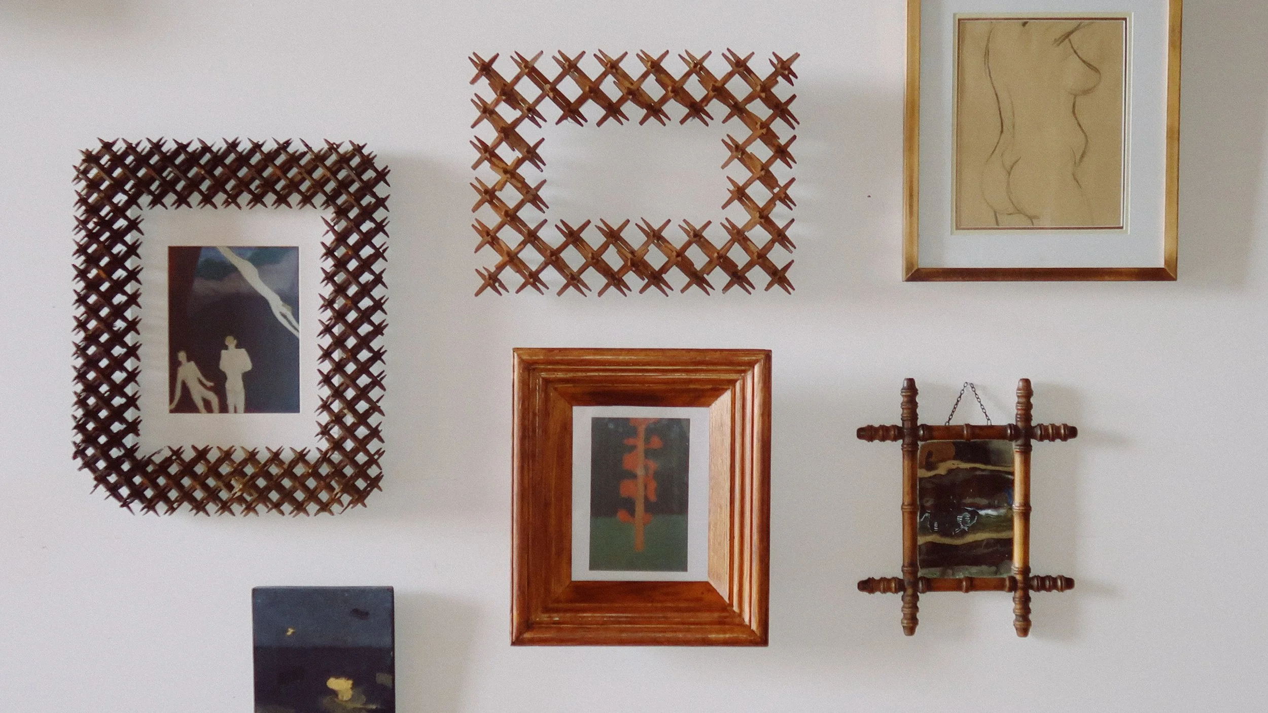 Collection of five framed artworks on a white wall, including geometric and abstract designs, with woven and wooden tramp art frames.