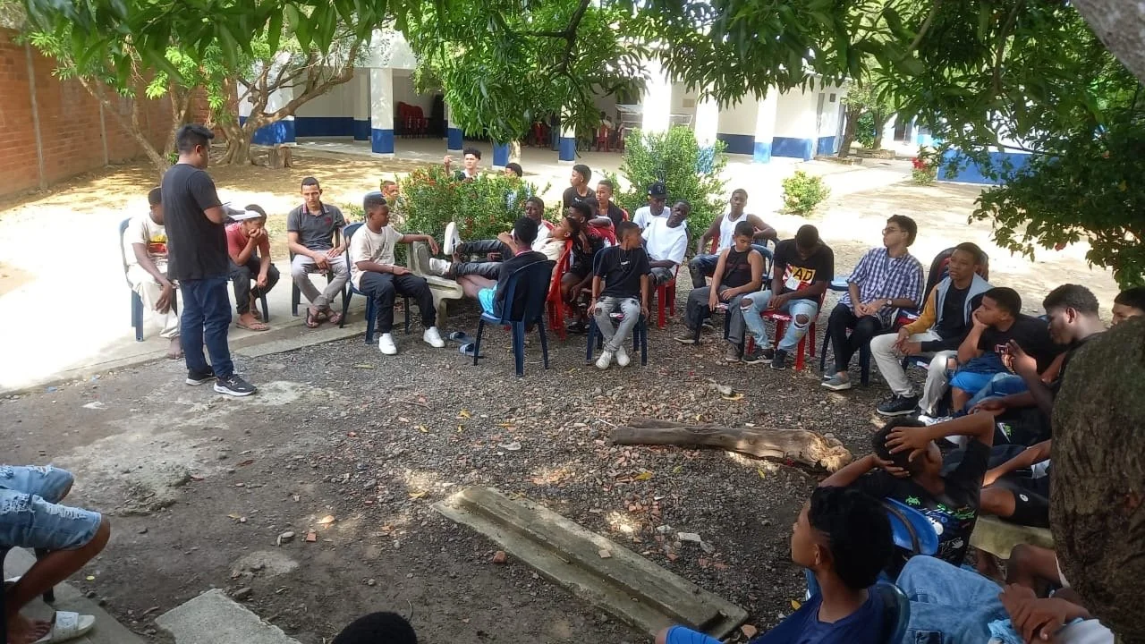 A group of young people sitting in a semi-circle outdoors, listening to a person standing and speaking. There are trees providing shade, and a building is visible in the background.