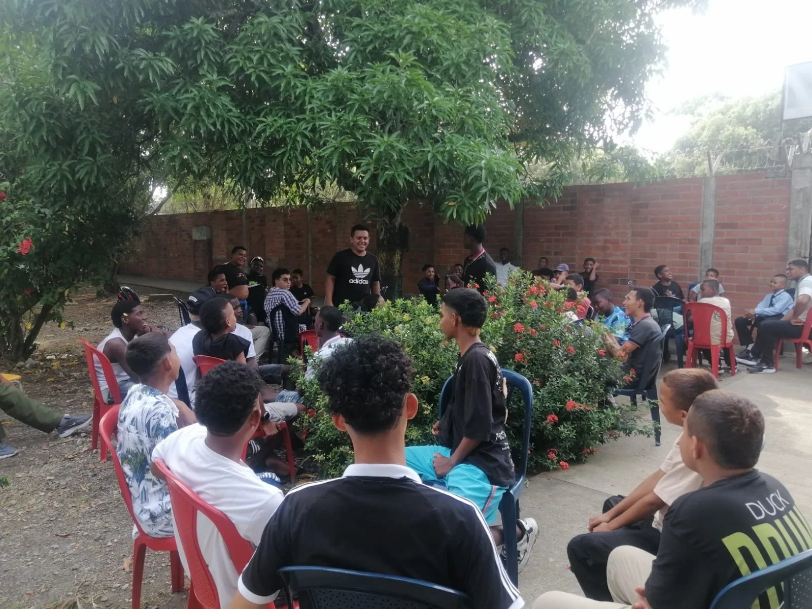 Group of young people gathered outdoors, some sitting on colorful chairs around a central bush, others standing, as they listen to a man speaking near a brick wall and lush green trees.