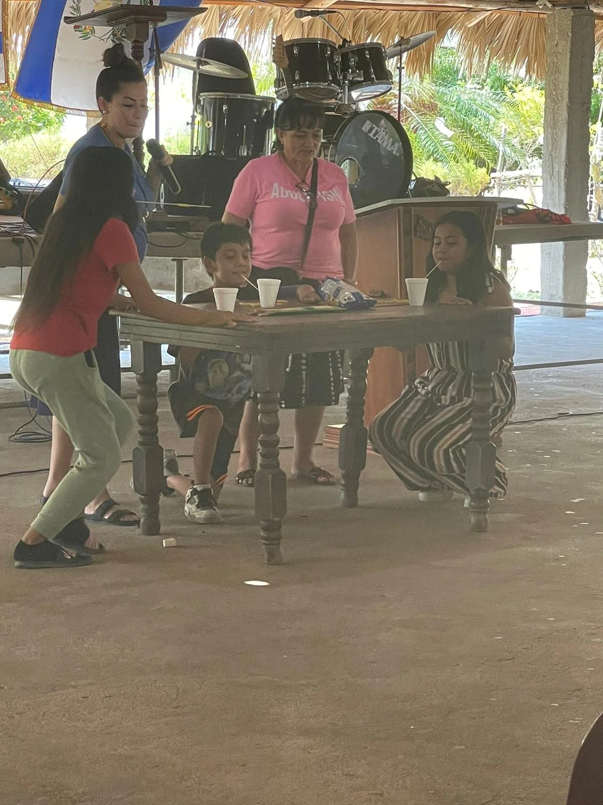 Three children and two women are participating in a game where they fish for prizes or items on a table inside a venue with a thatched roof. One woman appears to be helping, and there is musical equipment in the background.