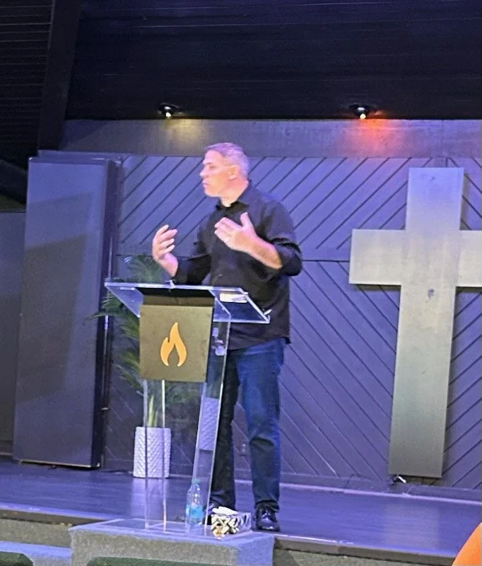 A man speaking at a church or religious venue, standing behind a clear lectern with a flame symbol, with a cross on the wall behind him.