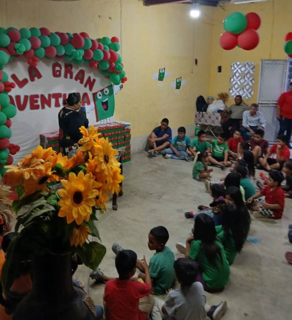 Children and adults sitting on the floor in a decorated room, listening to a woman speak. Decor includes red and green balloons and large sunflower bouquet. Wall decorations include cartoon character cutouts and a banner with red letters.