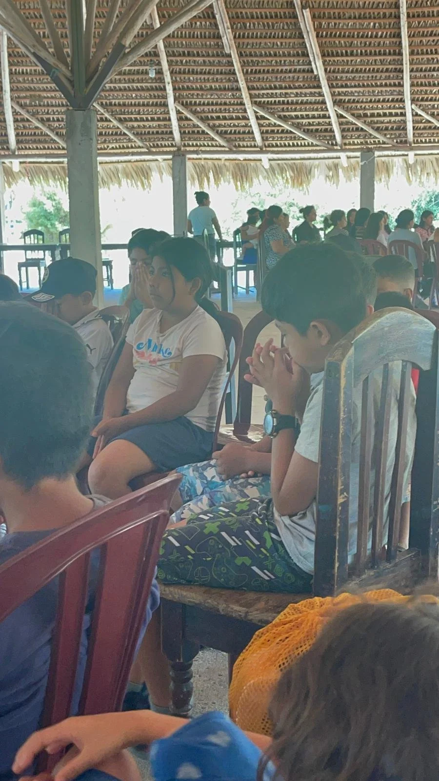 Children sitting on chairs inside a thatched-roof open-air structure, some with hands clasped and eyes closed, others looking around, with adults and more children in the background.