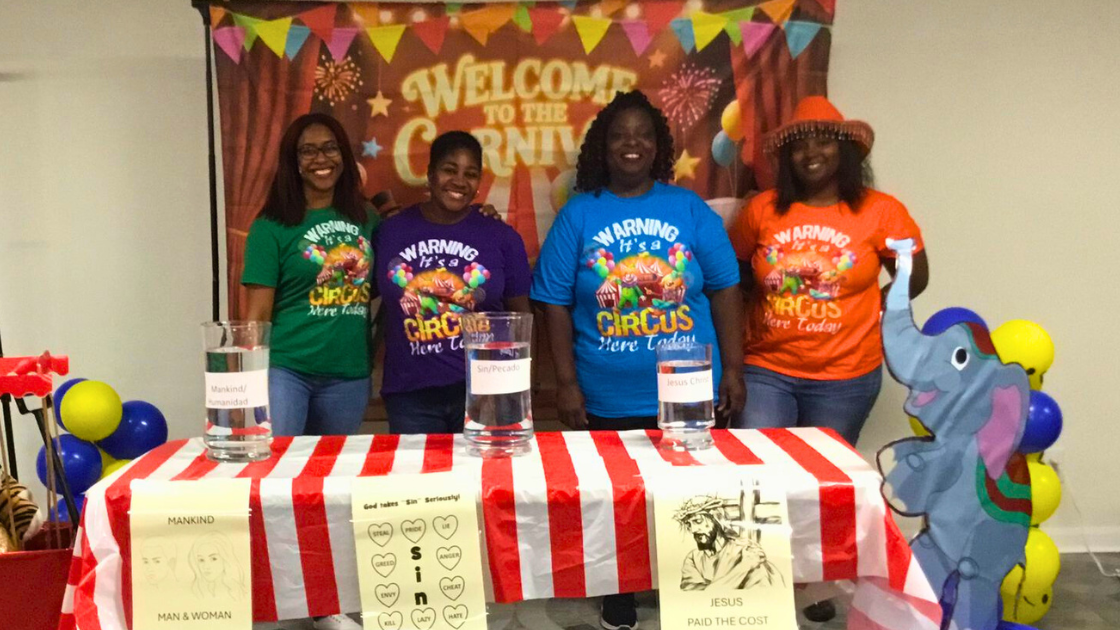 Four women stand behind a table decorated for a circus-themed event, with colorful balloons and a large cartoon elephant on the right. They are wearing bright T-shirts with circus warning messages. The background banner says 'Welcome to the Carnival.' The table has labels, posters, and jars of water.