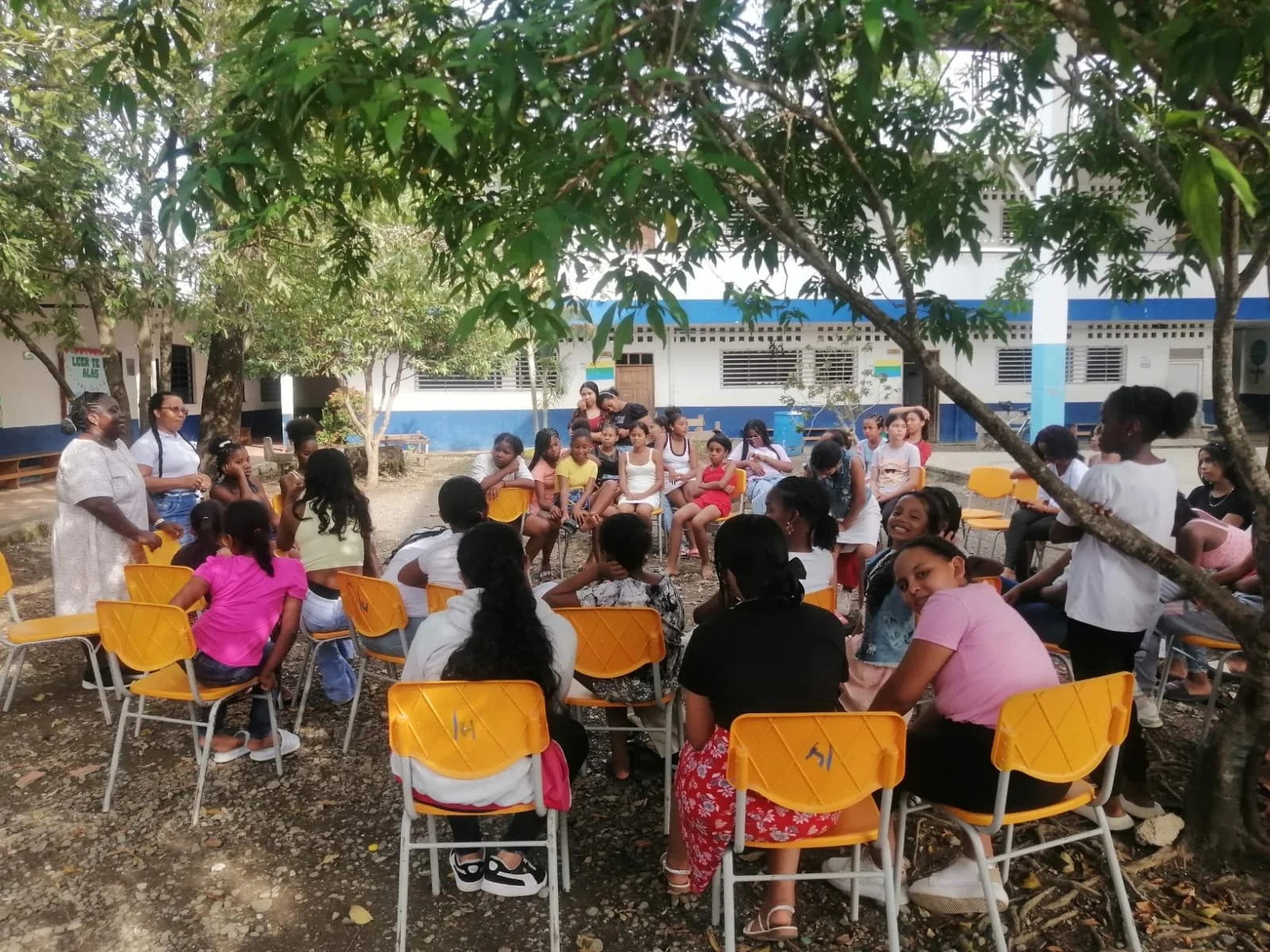 A group of children and a few adults gathered outdoors in a courtyard under trees, sitting on yellow chairs arranged in a circle, with some people standing and speaking.