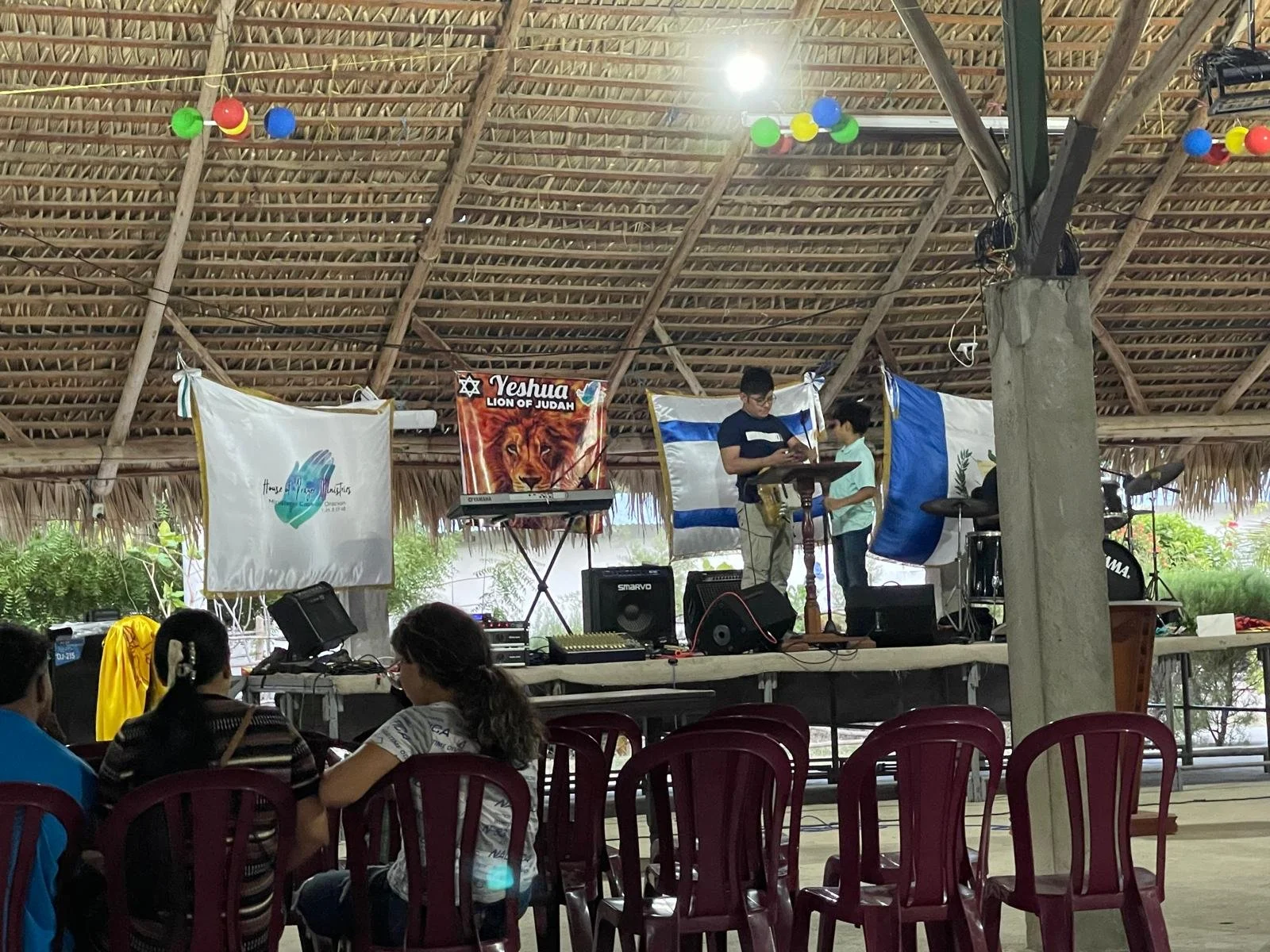 A stage set up under a thatched roof with banners and musical equipment, with a few people standing and sitting in front of it.