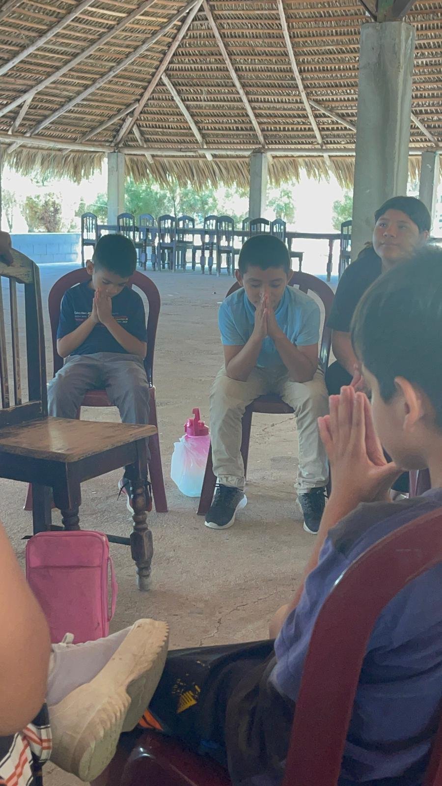 Children and a young man praying with hands folded in a semi-circle under a thatched roof structure.