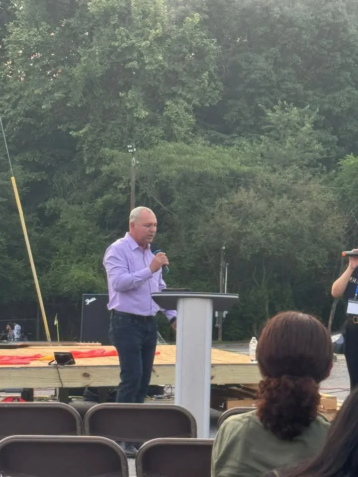 A man in a purple shirt speaking into a microphone at an outdoor event with a wooden stage, surrounded by trees, with a woman standing to his right and seated audience members in the foreground.