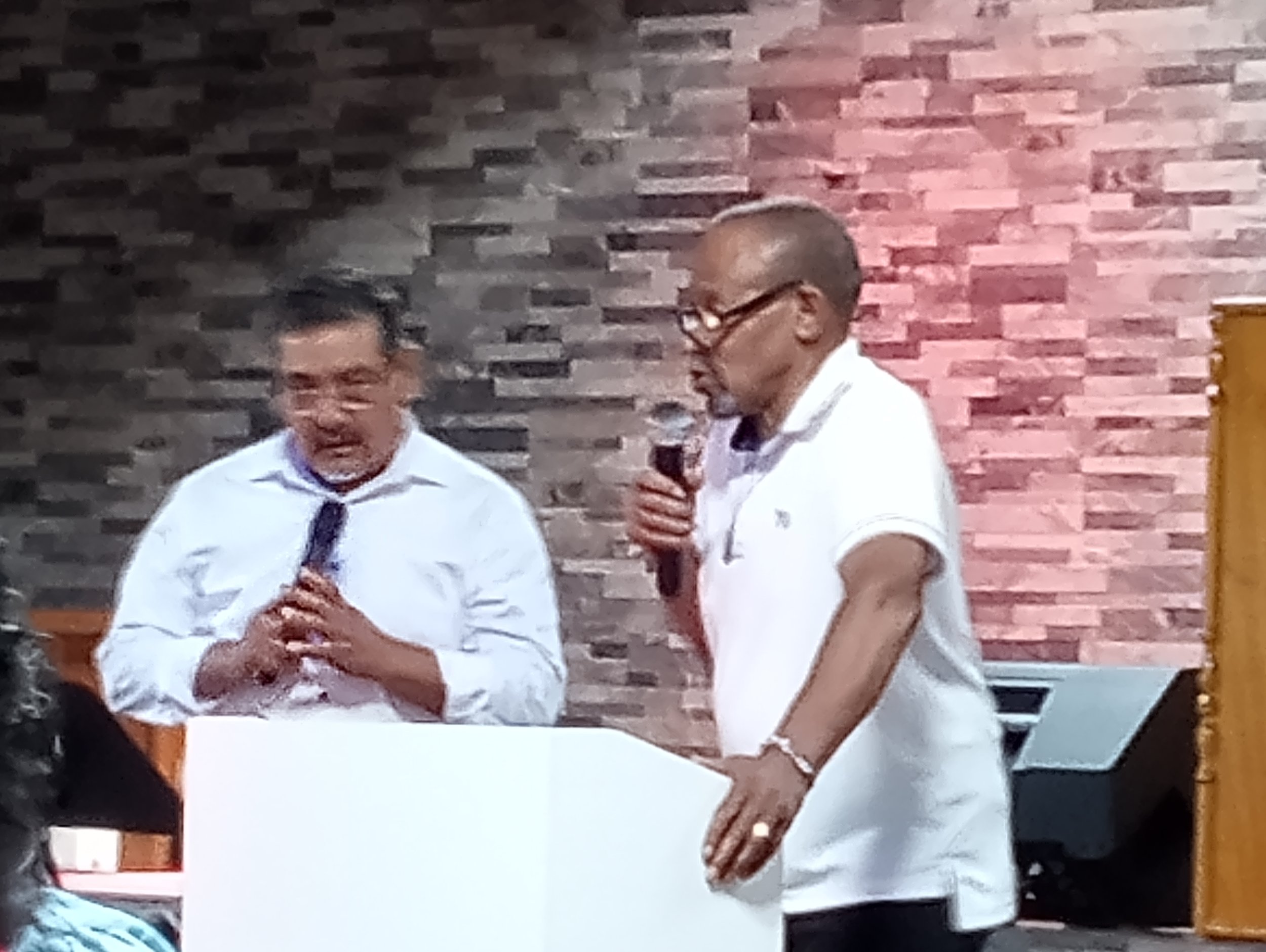 Two men speaking into microphones indoors, with a brick wall background, one with glasses and short hair, and the other with glasses and a shaved head.