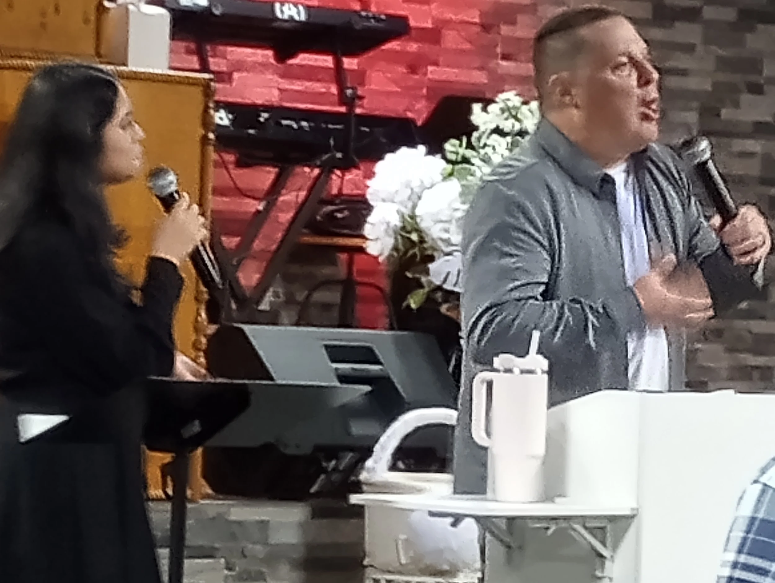 A man and woman holding microphones speaking at an indoor event with flowers and musical instruments in the background.