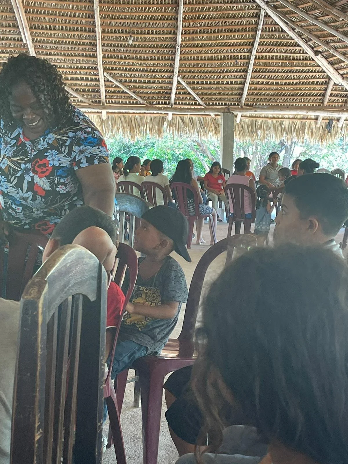 People gathered indoors under a thatched roof, sitting on chairs, engaged in conversation, with some children and adults present.
