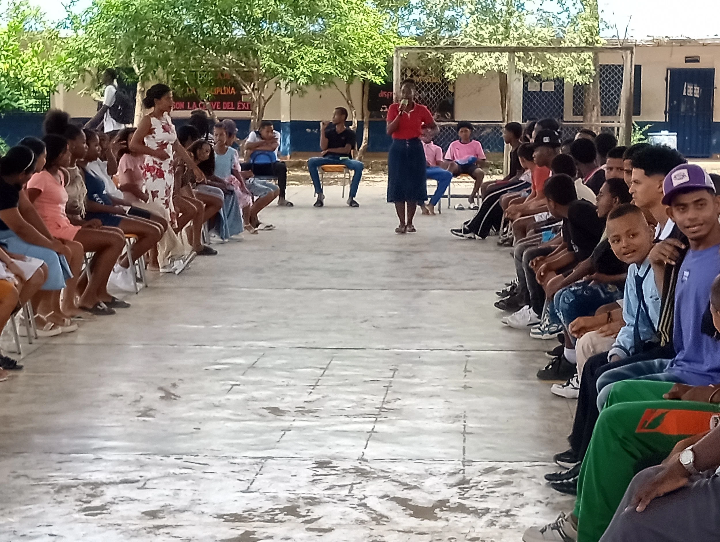 Children and teenagers seated in two long rows facing each other outdoors, with a woman standing and speaking in the middle. Another woman is seated in the background, under a tree, with a few more children sitting behind her. The setting appears to be a school or community event on a concrete courtyard with trees and buildings in the background.