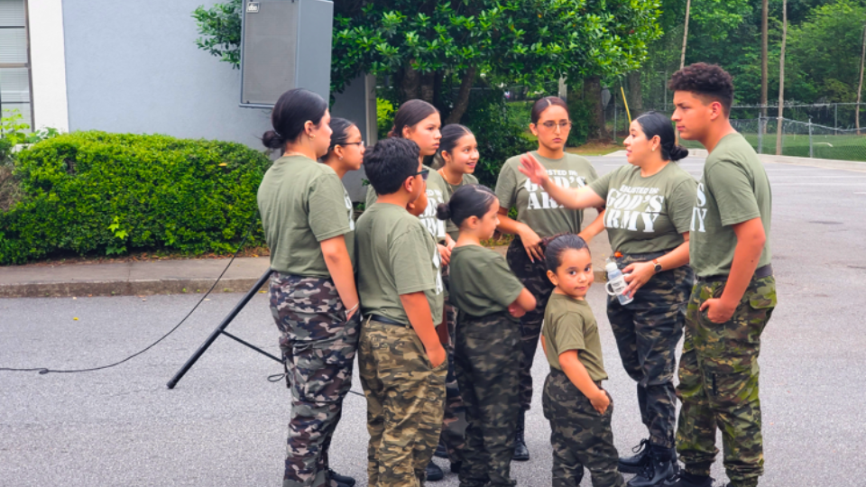Group of young girls and a young man in military camo clothing, standing outdoors on pavement, engaged in conversation, some wearing glasses, with green bushes and trees in the background.